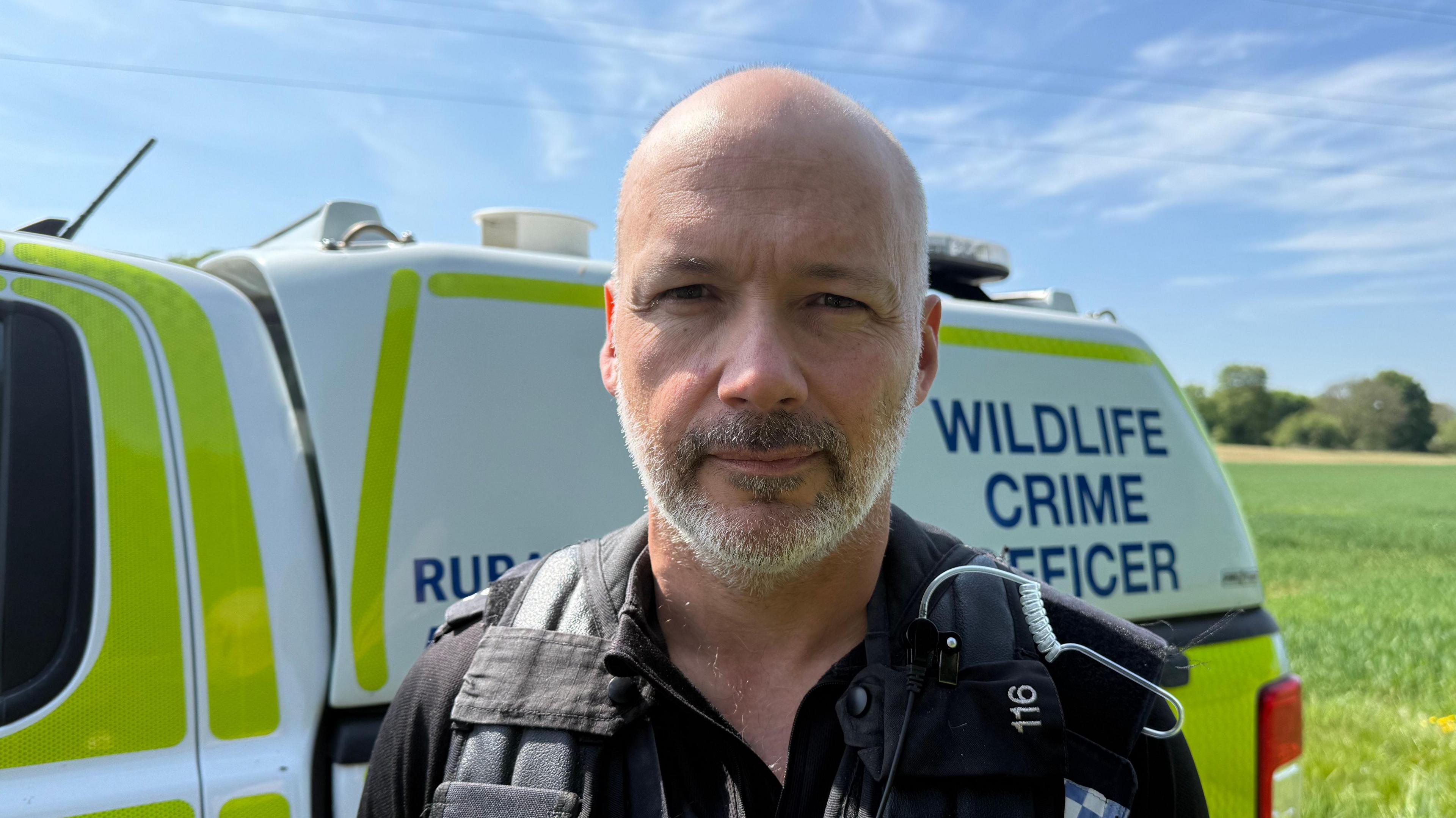 Head and shoulders shot of Det Con Aaron Flint. He has a short beard and is standing in front of a rural crime patrol car. There is greenery in the background.