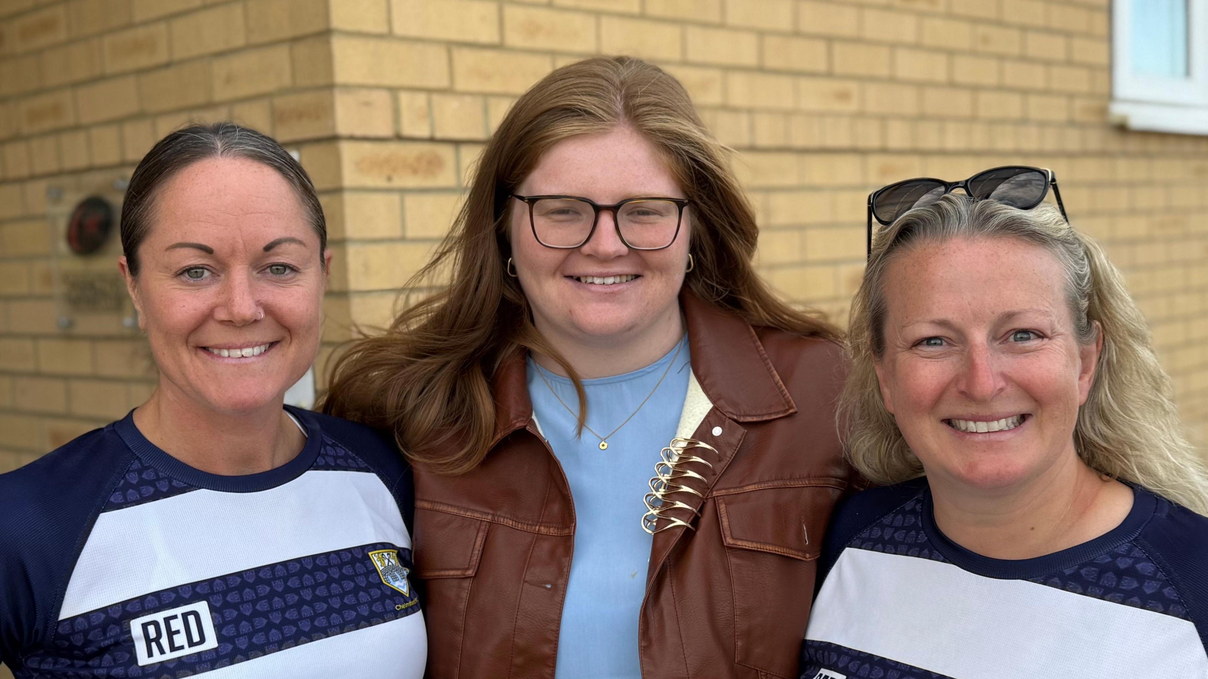 Three women smiling straight at the camera while standing in front of a brick wall. Two of them are in rugby kit and the other is wearing a blue top and brown coat.