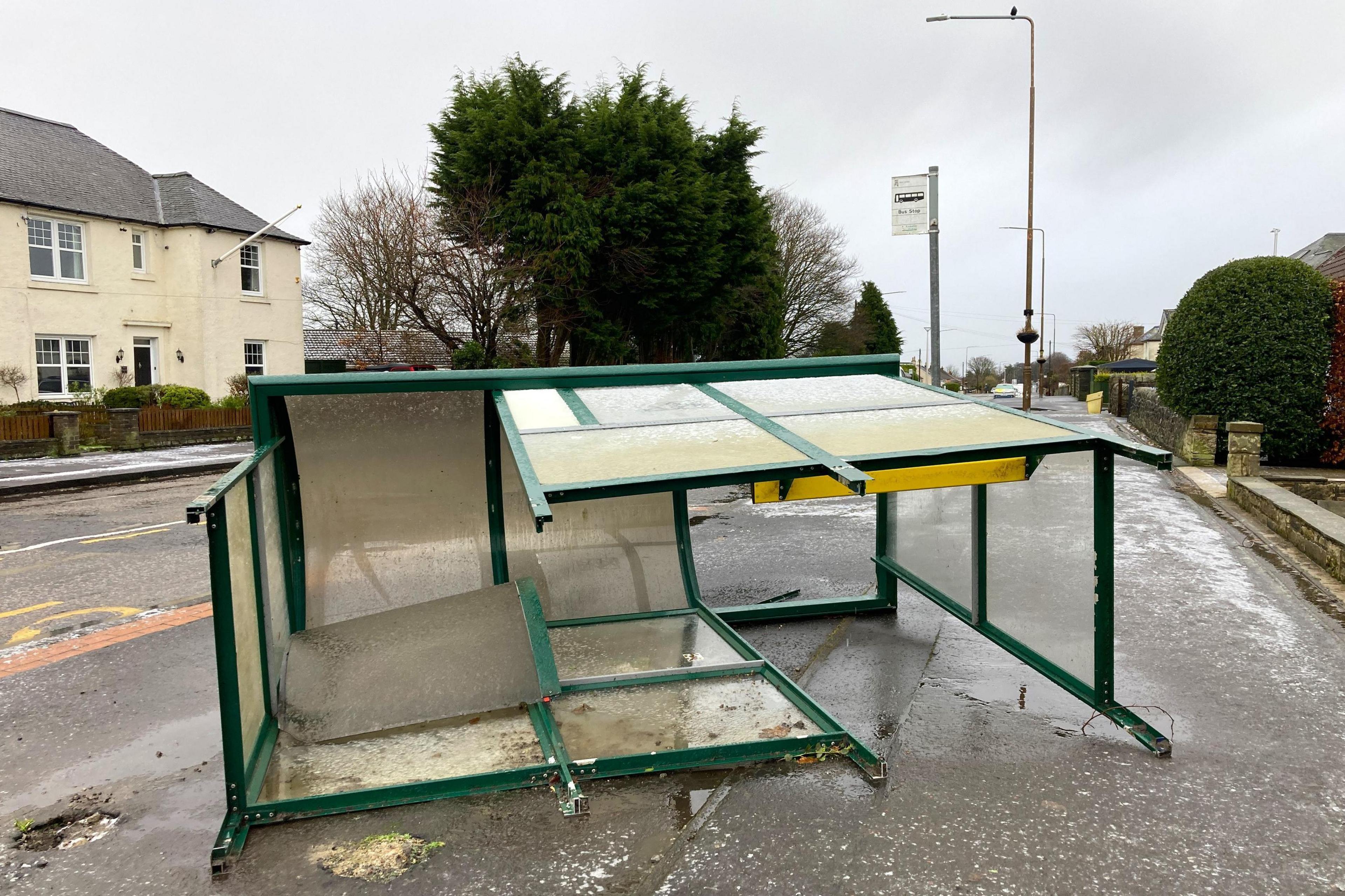 A smashed up green and glass bus stop lies on its side on a pavement. There are houses in the background.