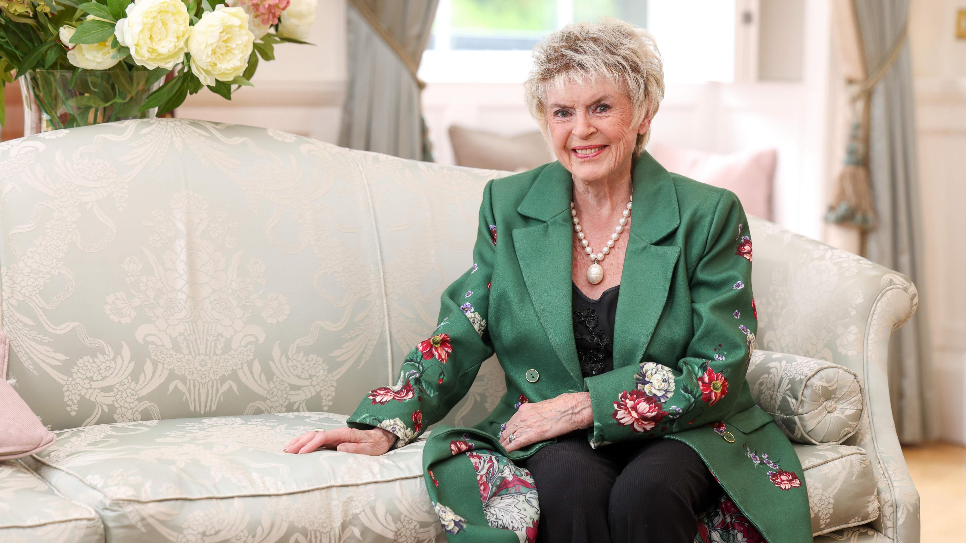 Gloria Hunniford with short blonde hair, wearing a long green jacket, a black top and black trousers, is sitting on a cream sofa. She is smiling at the camera. 