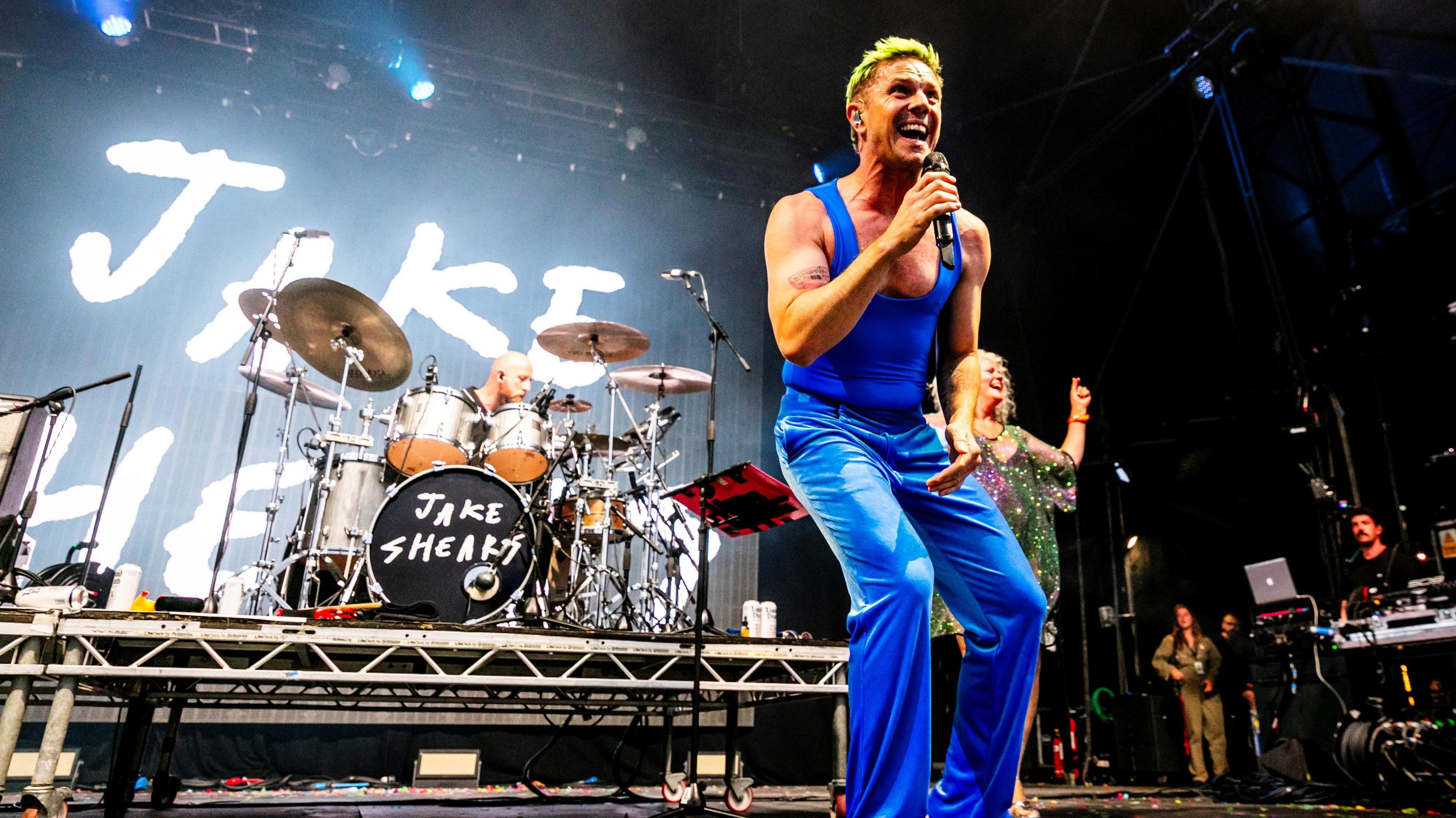 Jake Shears, dressed in a blue outfit, sings into the microphone while performing on stage