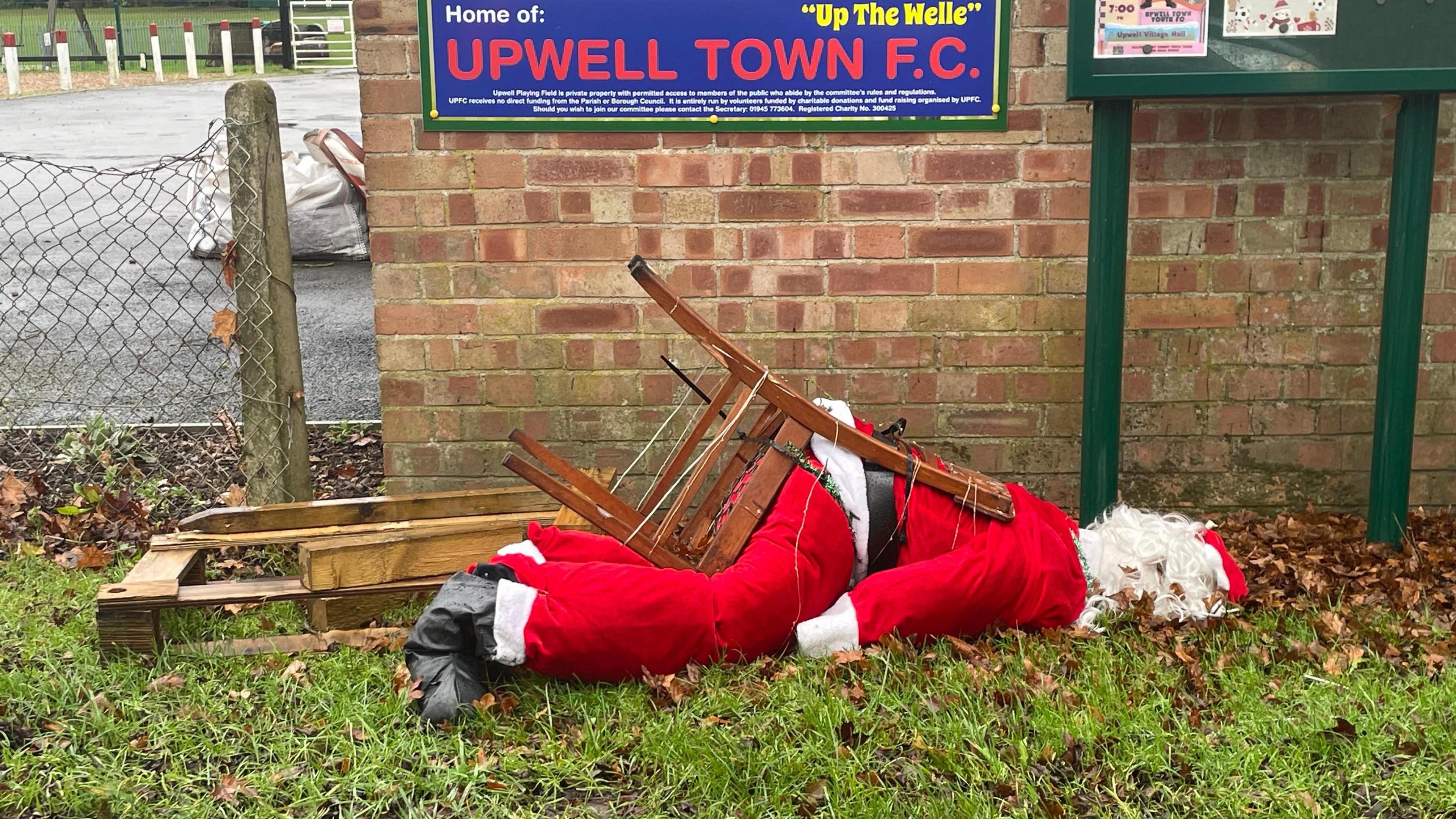 A Father Christmas on a wooden chair was sat outside a football club next to a sign and in front of a wall. However, the figure has fallen over on its side due to the wind.