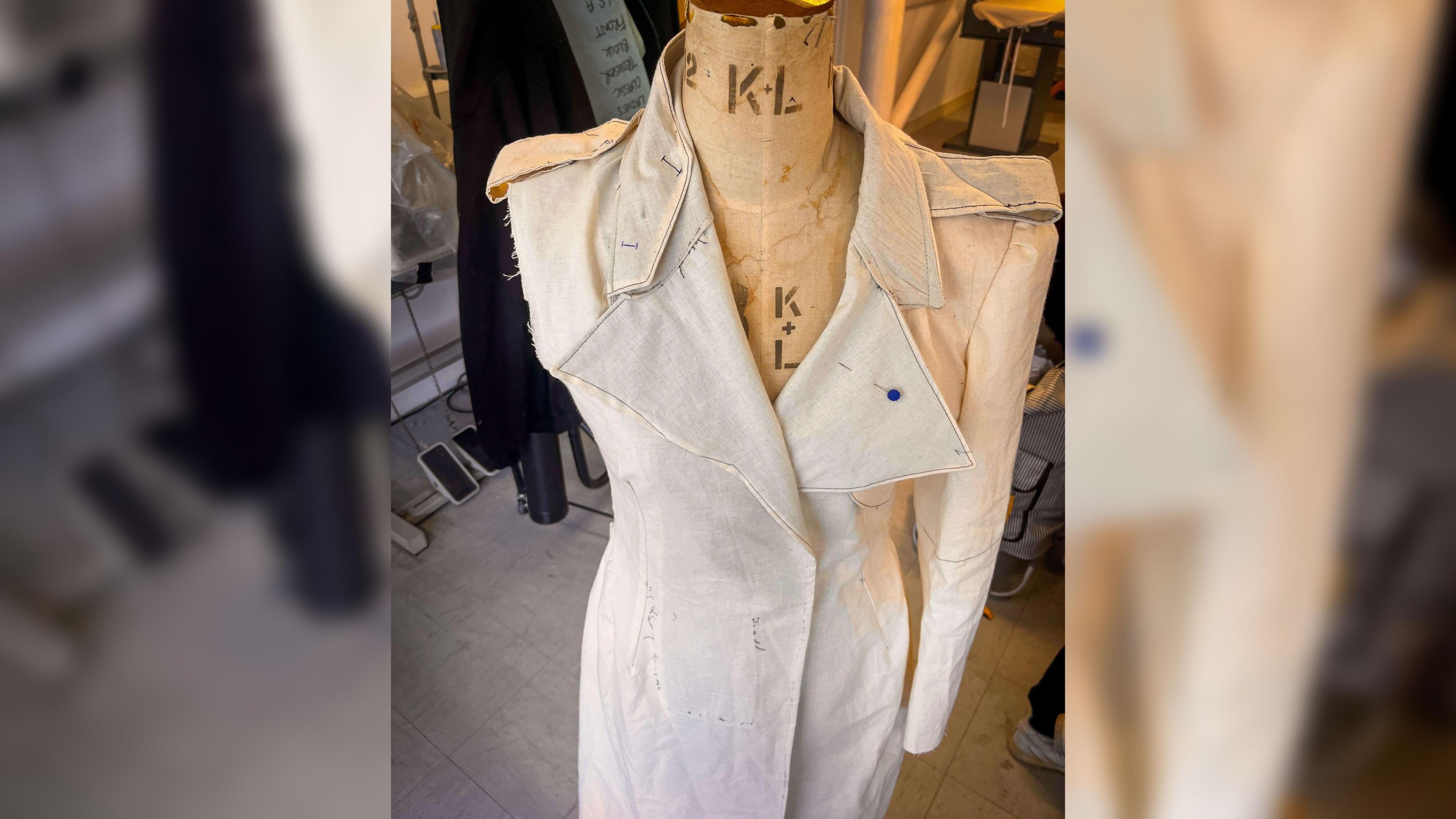 A mannequin with a partially-finished beige coat on it. There is one arm missing and a pin in the collar