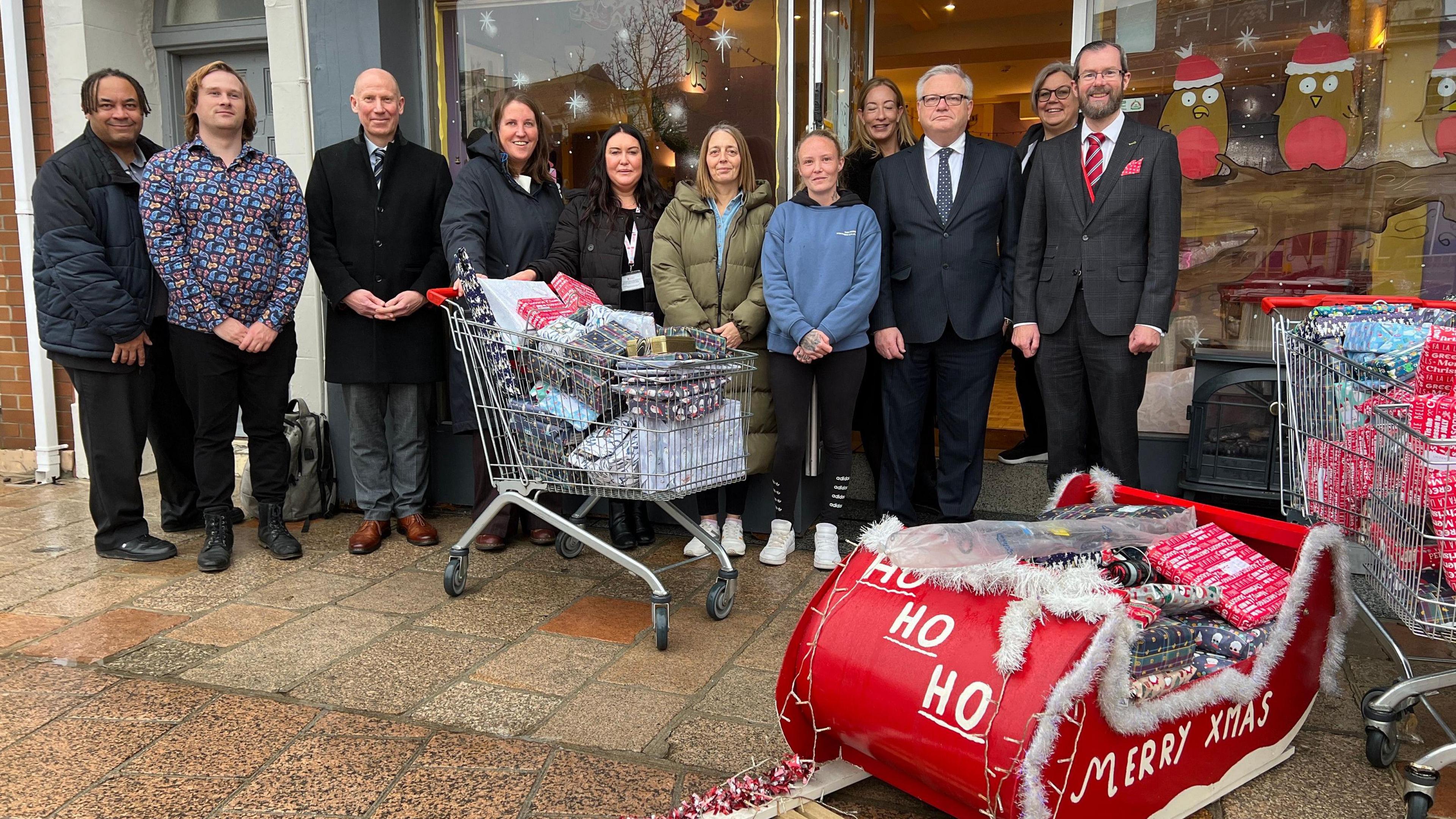 A group of people stand outside a cafe and presents are packed into trollies and a small red sledge which has ho ho ho Merry Xmas written in white on it.