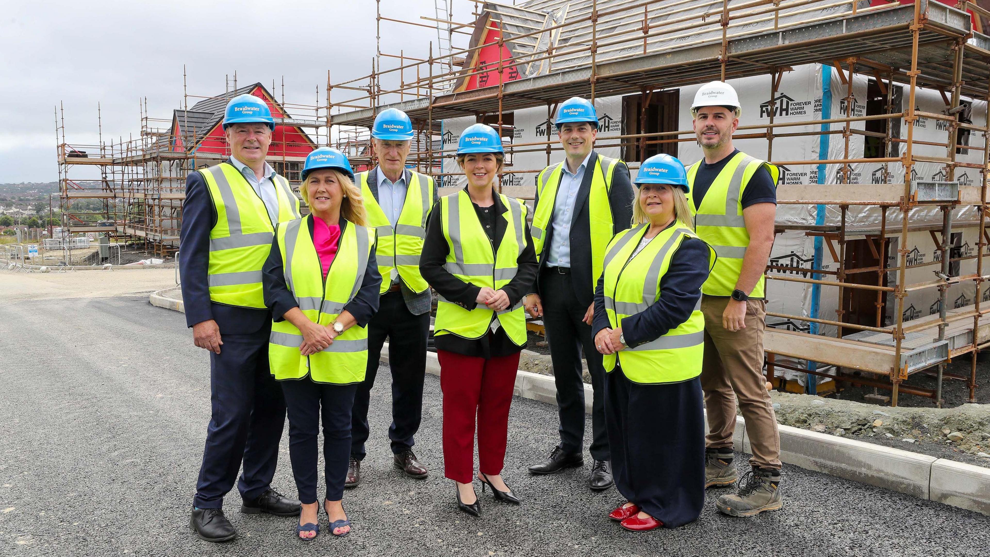 A group of seven people, all in hard hats and yellow high viz jackets stand in front of a house that is under construction. the house is covered by scaffolding.