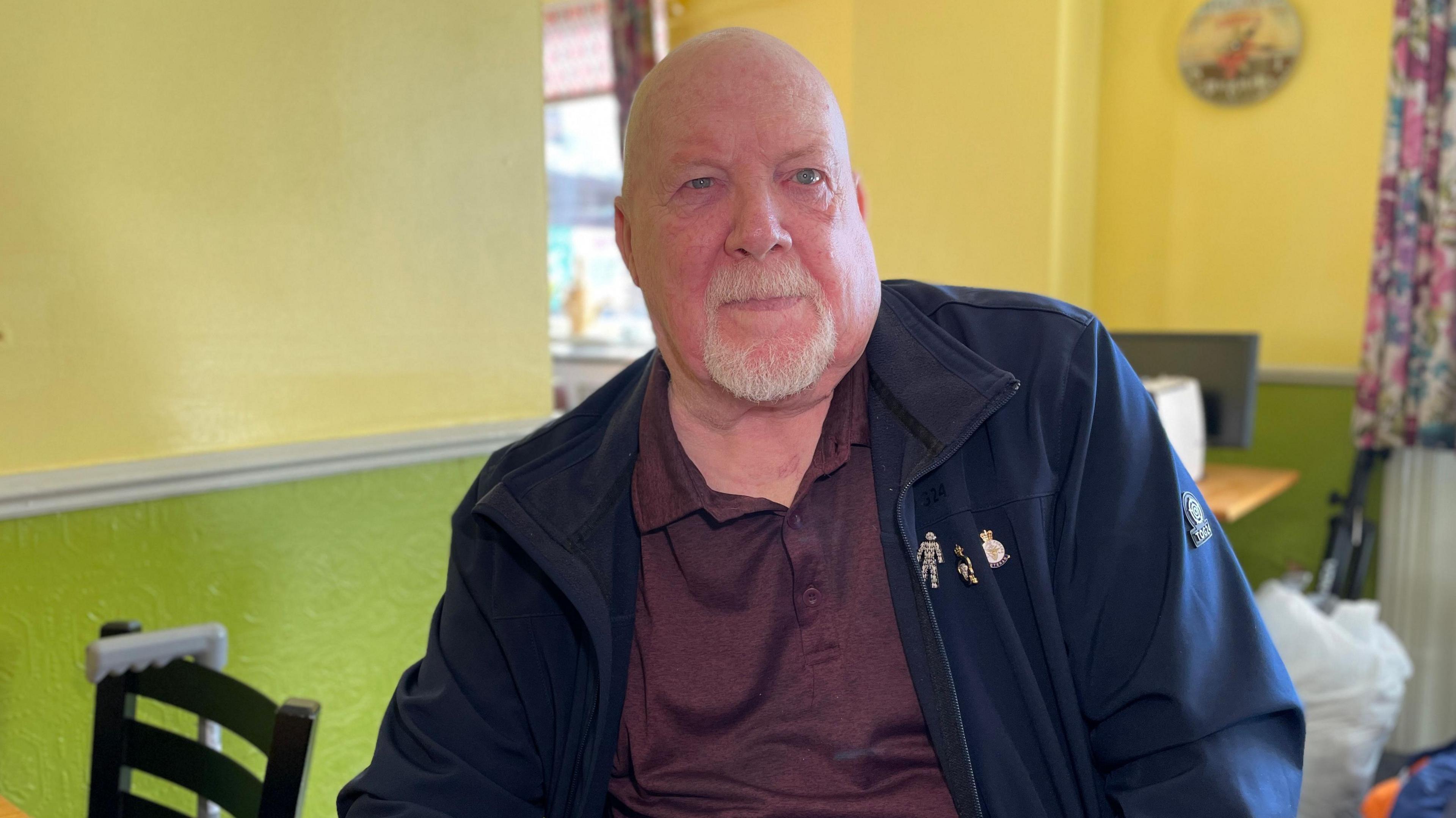 A close up picture of Ken Warnes. He is sat on a chair in a cafe and is wearing a navy blue jacket with three badges on and burgundy t-shirt. He has a bald head and a light grey beard. The walls of the cafe are painted yellow and green. 