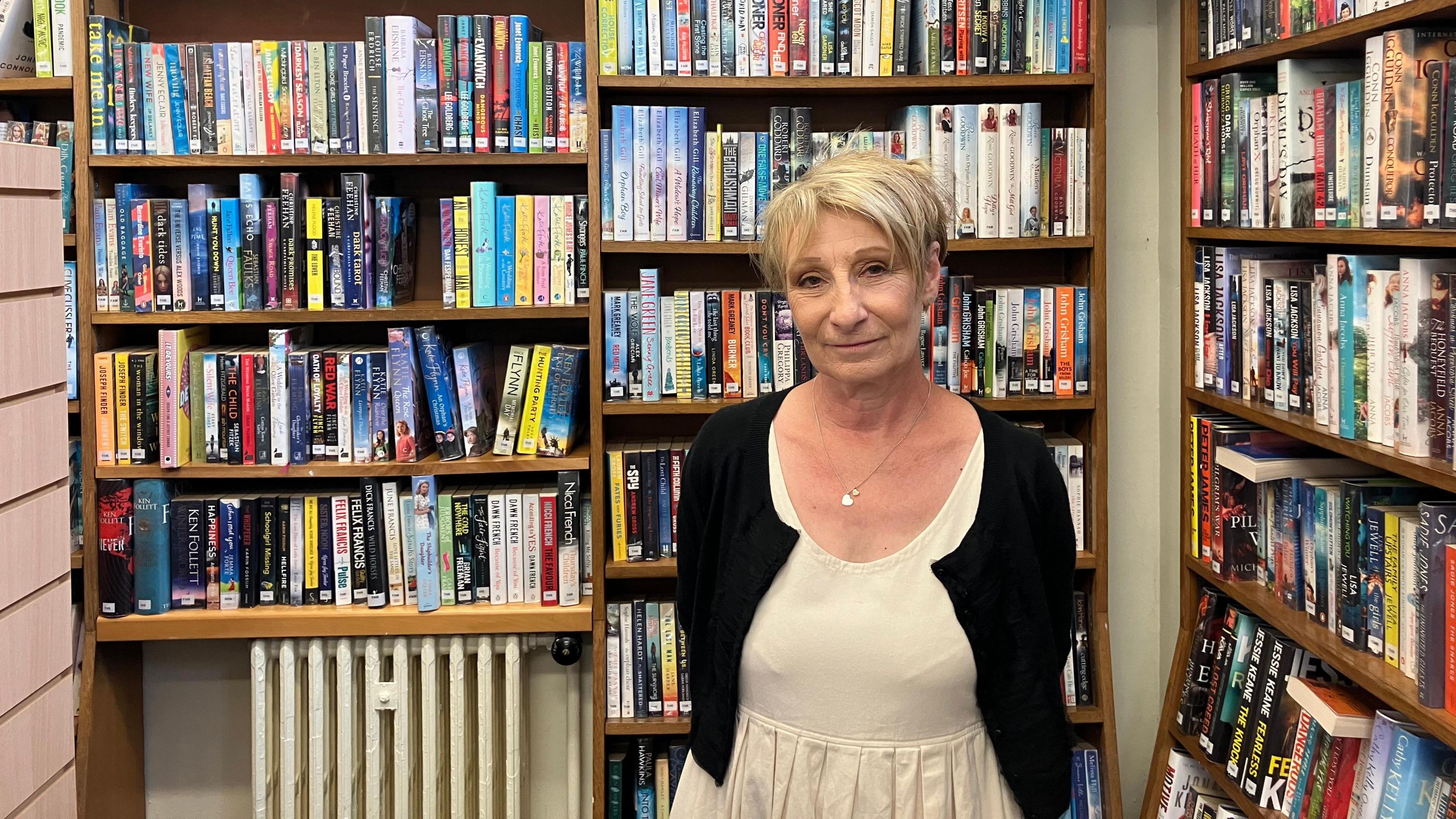 Jennie Storey, wearing a white dress and dark cardigan, stands in front of a bookshelf in Woodston Library, Peterborough 