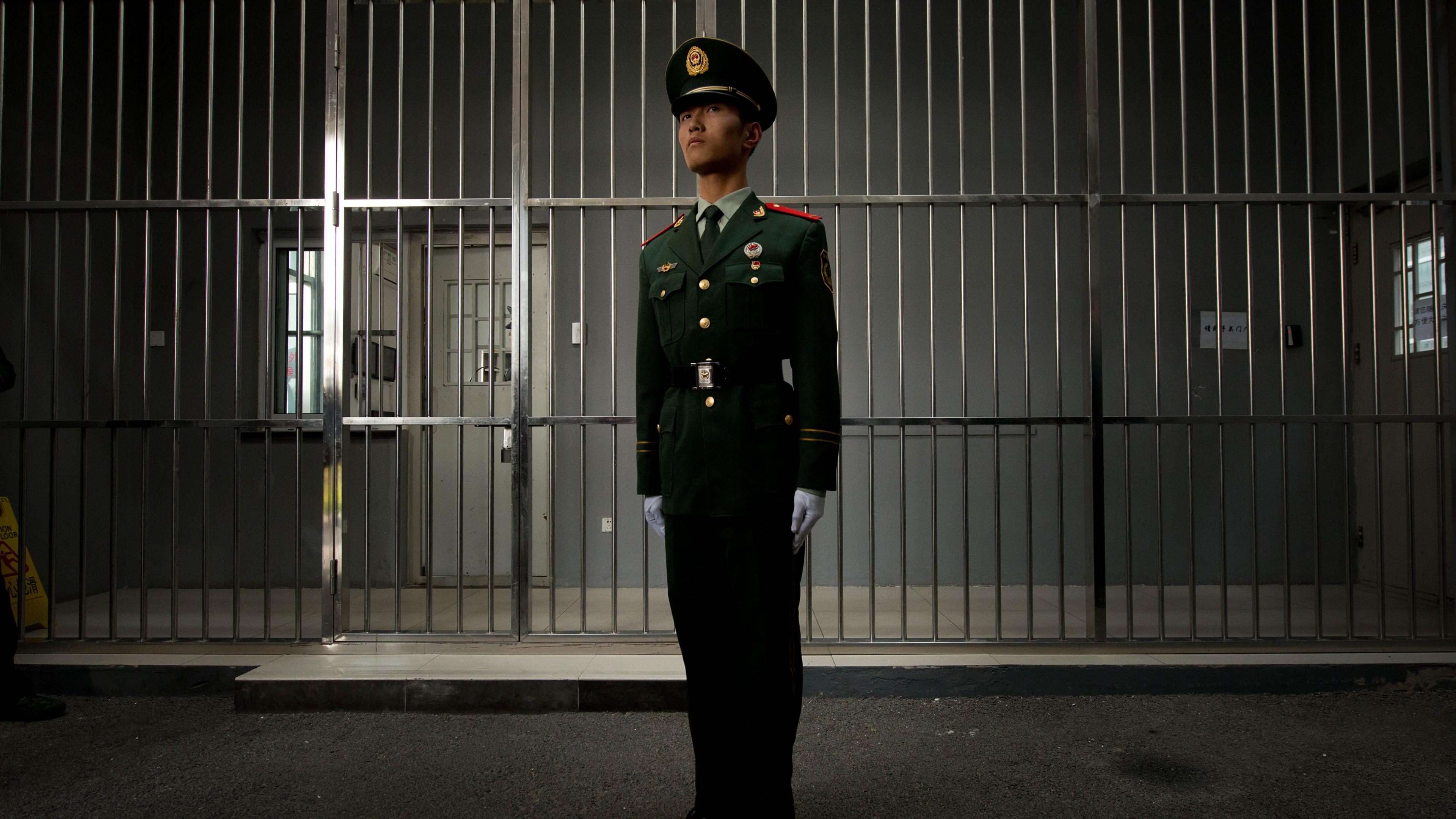 A paramilitary guard stands straight in his green uniform with white gloves in front of a wall of bars. Behind that is a corridor, with a white door and grey walls