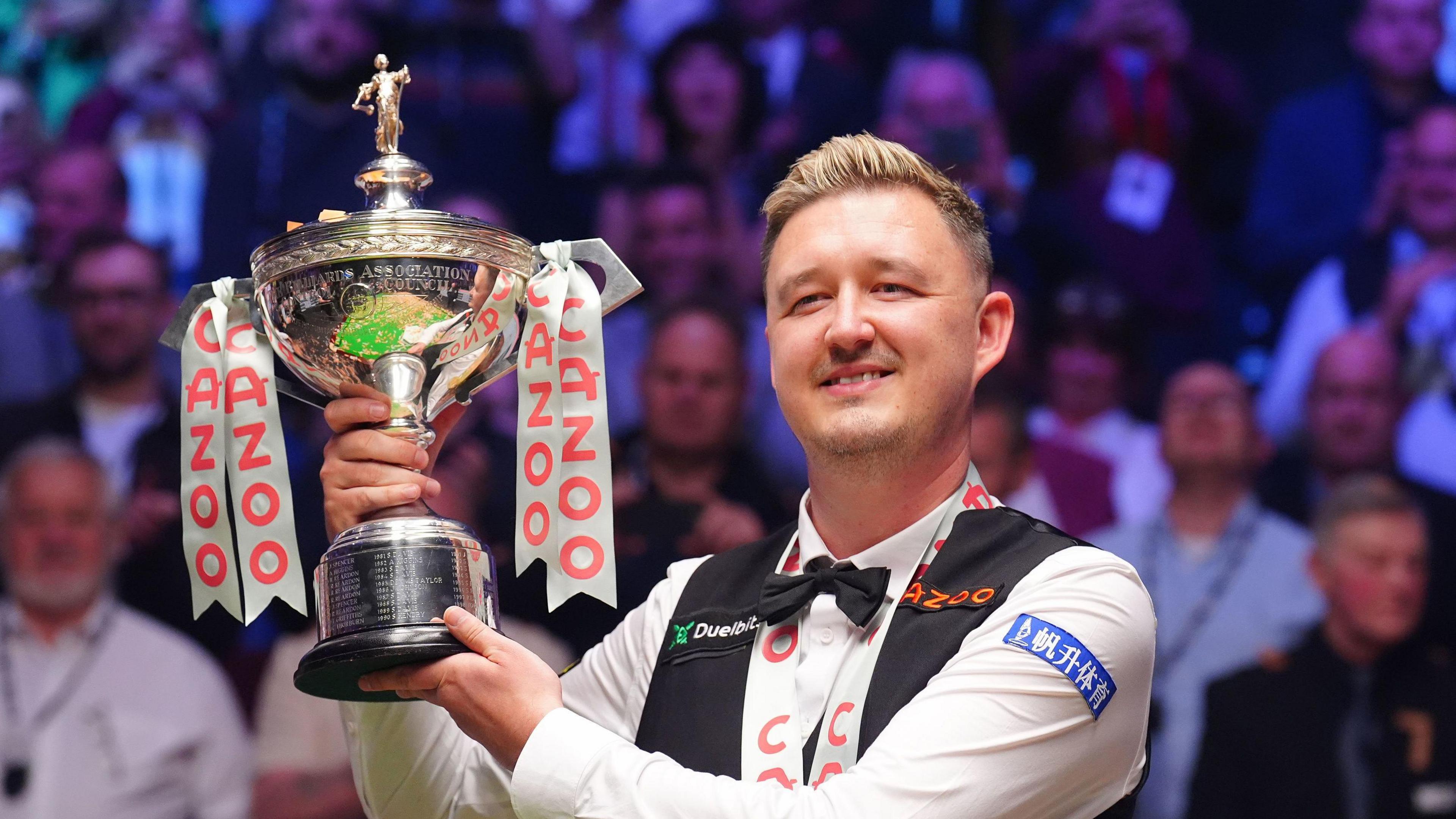 Kyren Wilson with short blonde hair wearing a black waistcoat and bowtie and white shirt with some logos printed on it. He is smiling and holding a large silver trophy.  There is an audience seated behind him.