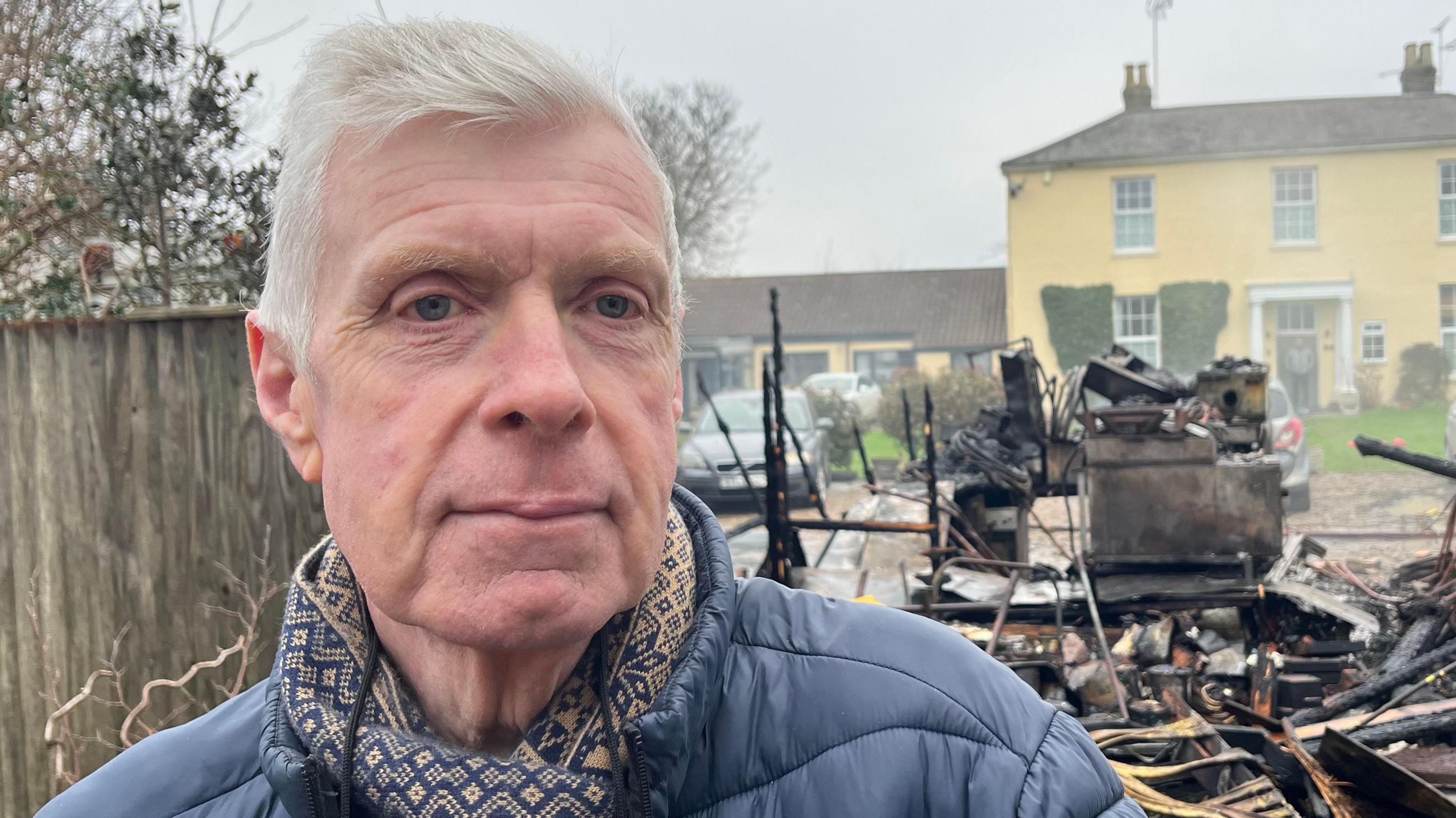 Ron Anderson looking into the camera while wearing a patterned scarf and blue coat. He is stood in front of burnt remains, with a yellow house on the opposite side of the street.