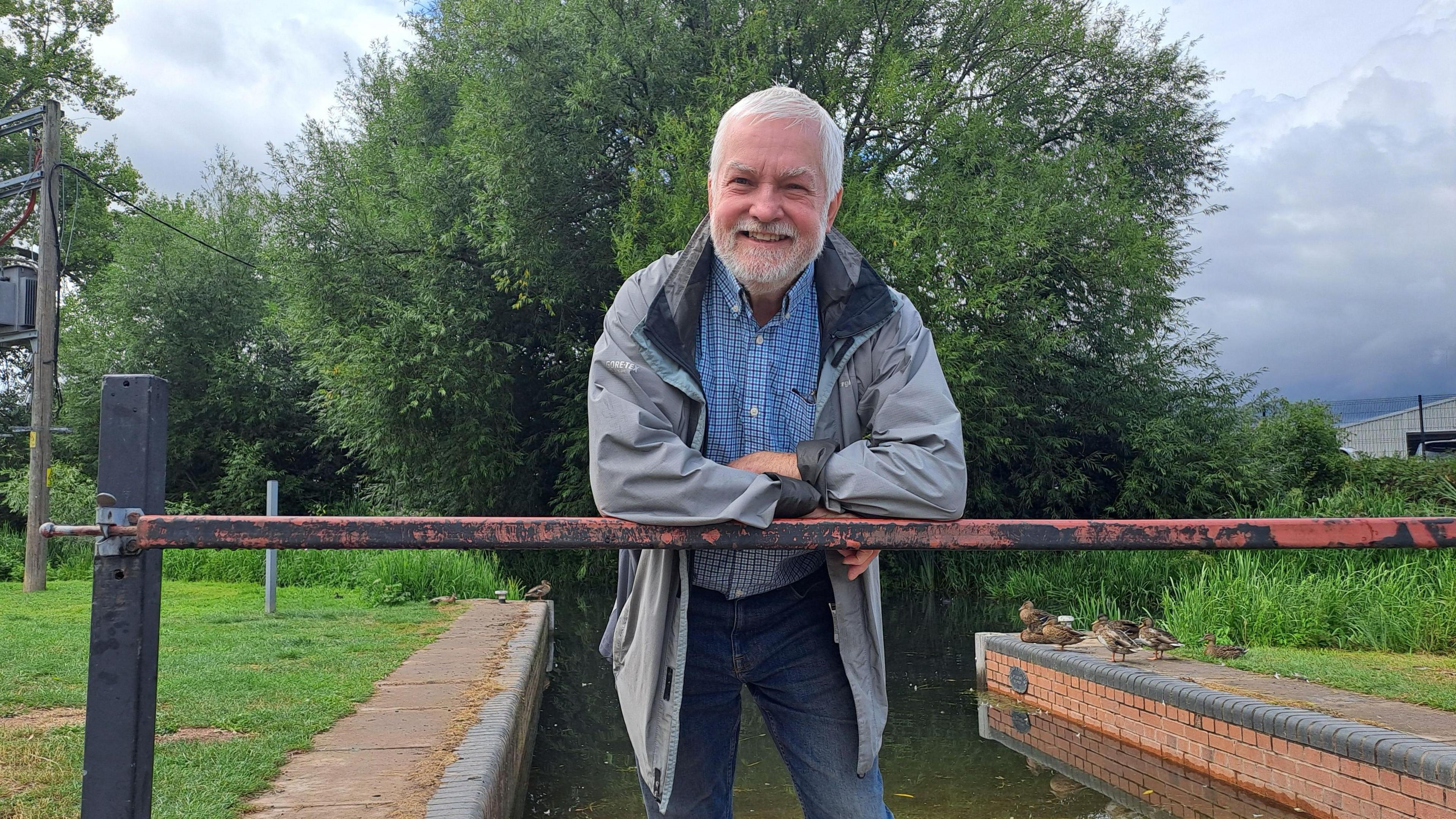 A man with short white hair leaning on a metal bar over a canal. He is smiling and has white facial hair. He is wearing a blue shirt and grey coat. There are trees and green grass surrounding the canal, with a group of ducks sitting alongside it