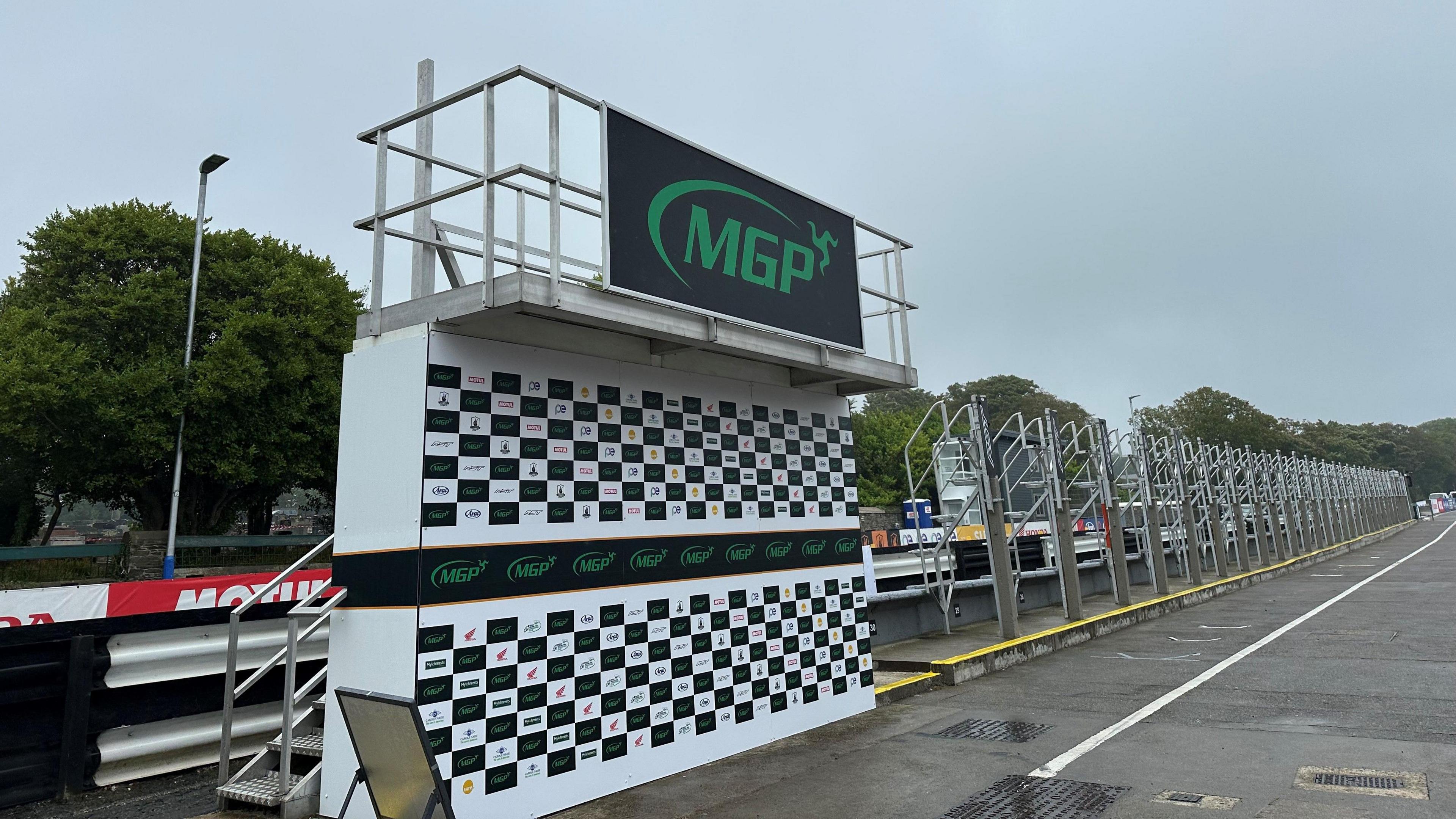 The Manx Grand Prix podium in and empty pit lane that is wet with rain. The podium has a chequered base, with a large sign with the MGP logo on it.