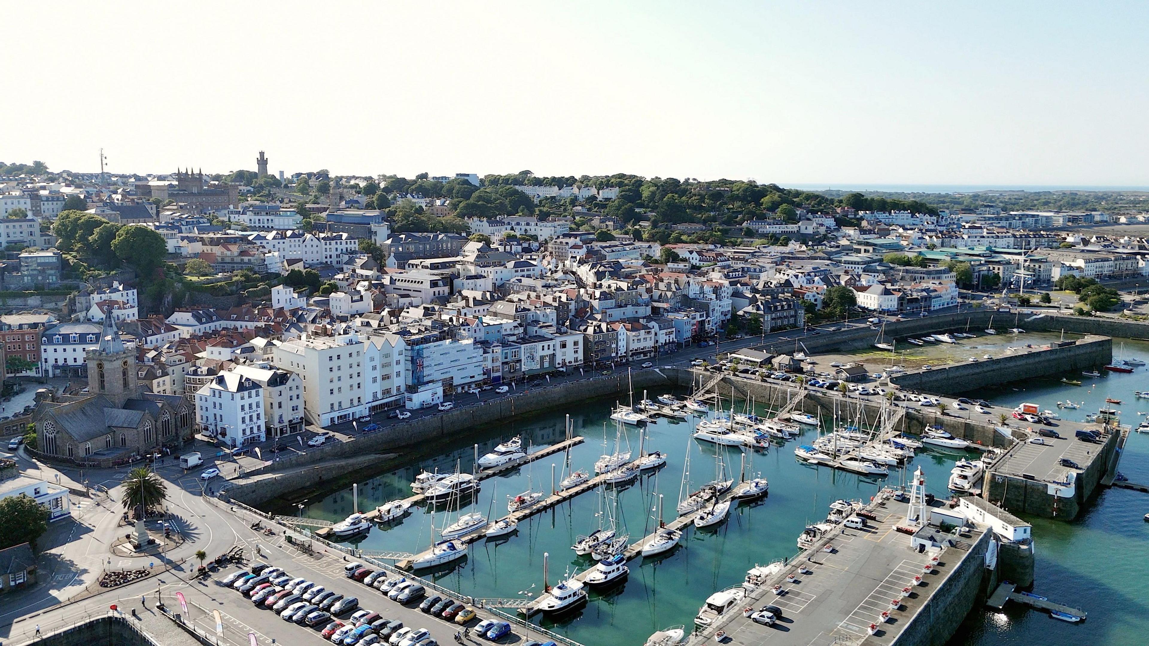 An aerial shot of St Peter Port in Guernsey. It shows boats in the harbour and a road with parked cars. Buildings can be seen in the background. 
