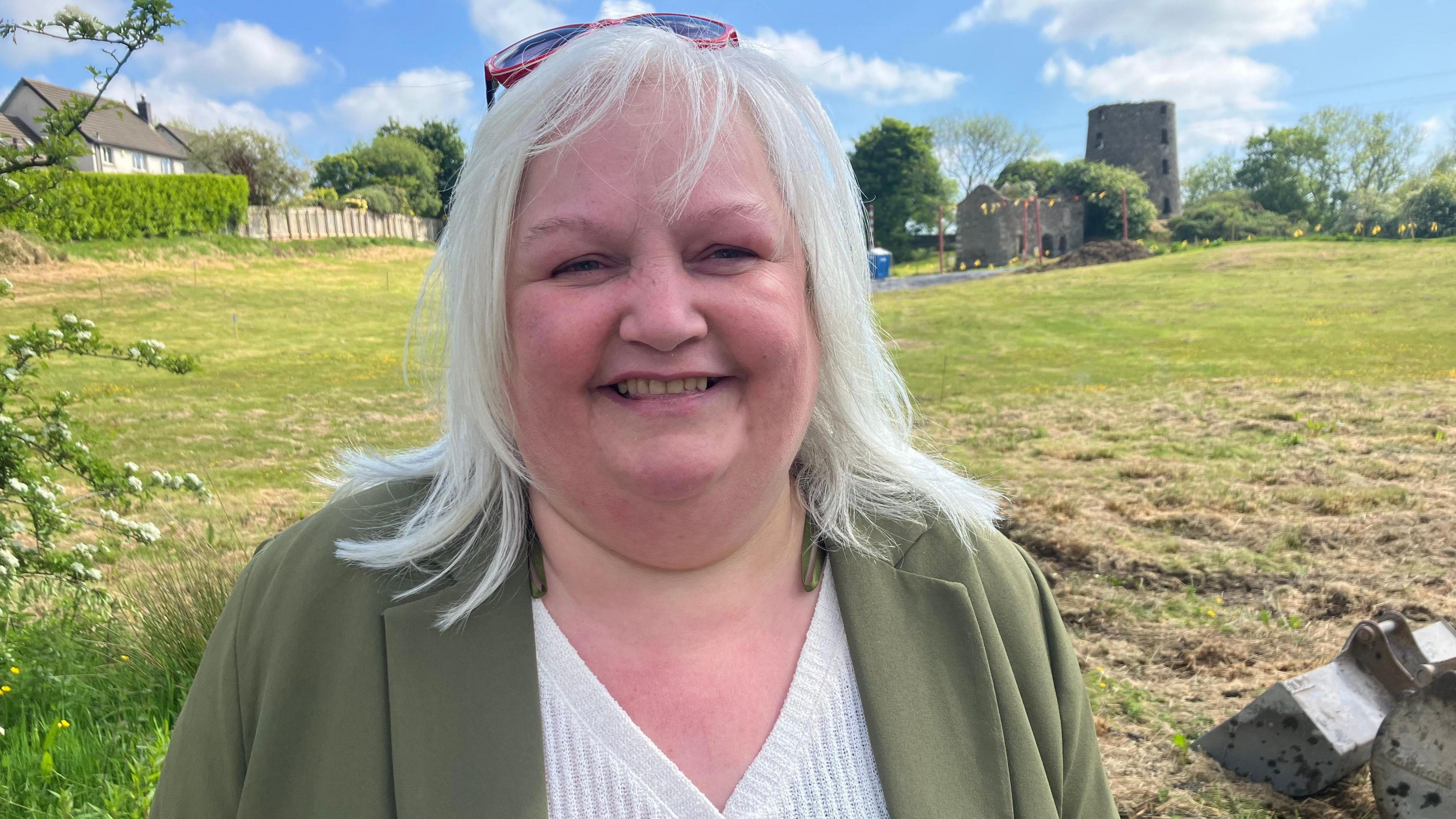 A woman with light coloured medium length hair stands in a field with ruins of a castle in the background. The sky is blue, and she is wearing a green blazer with white shirt underneath.