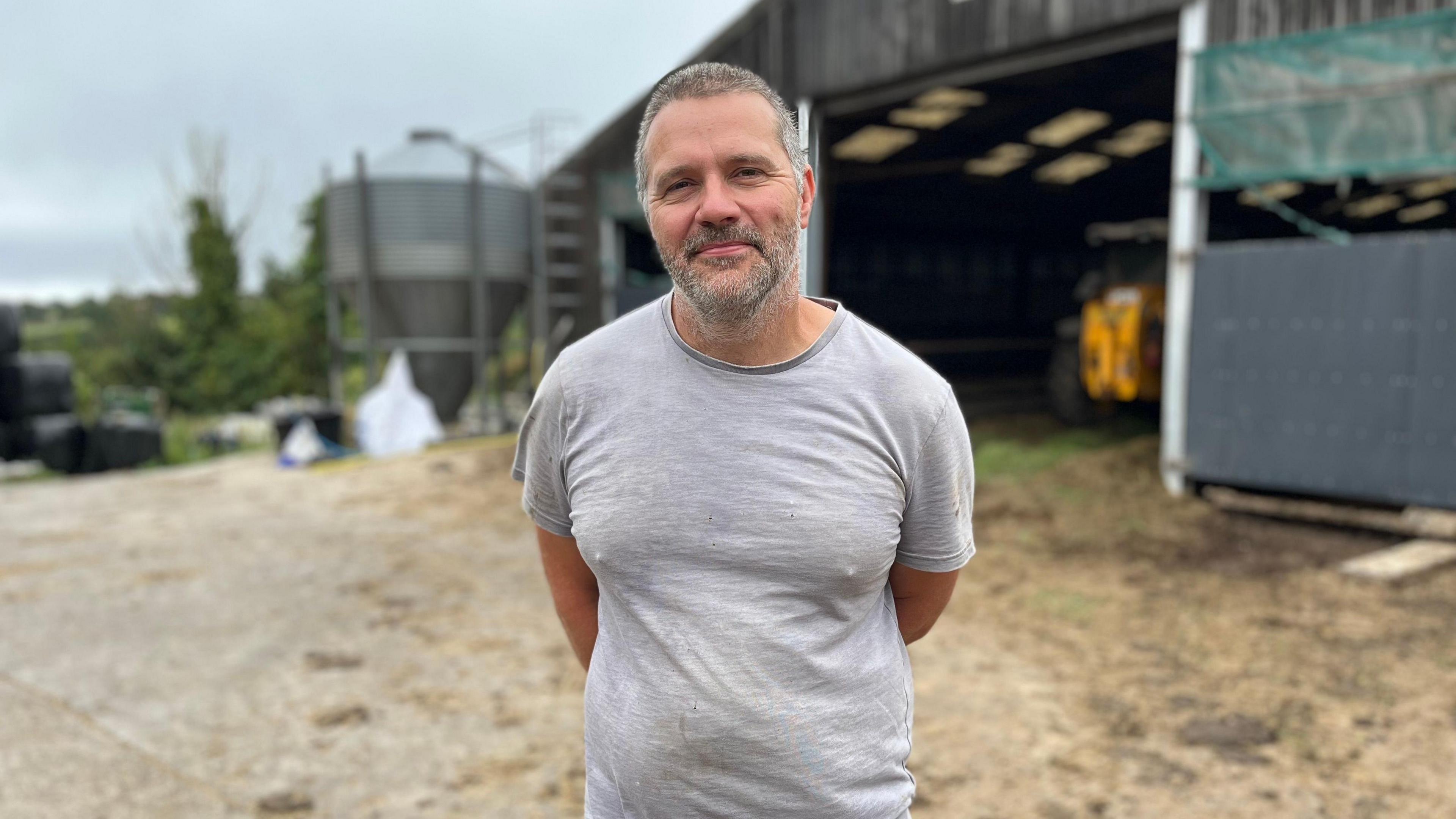 A man with short grey hair and a beard in a grey T shirt stands in front of a large wooden barn.