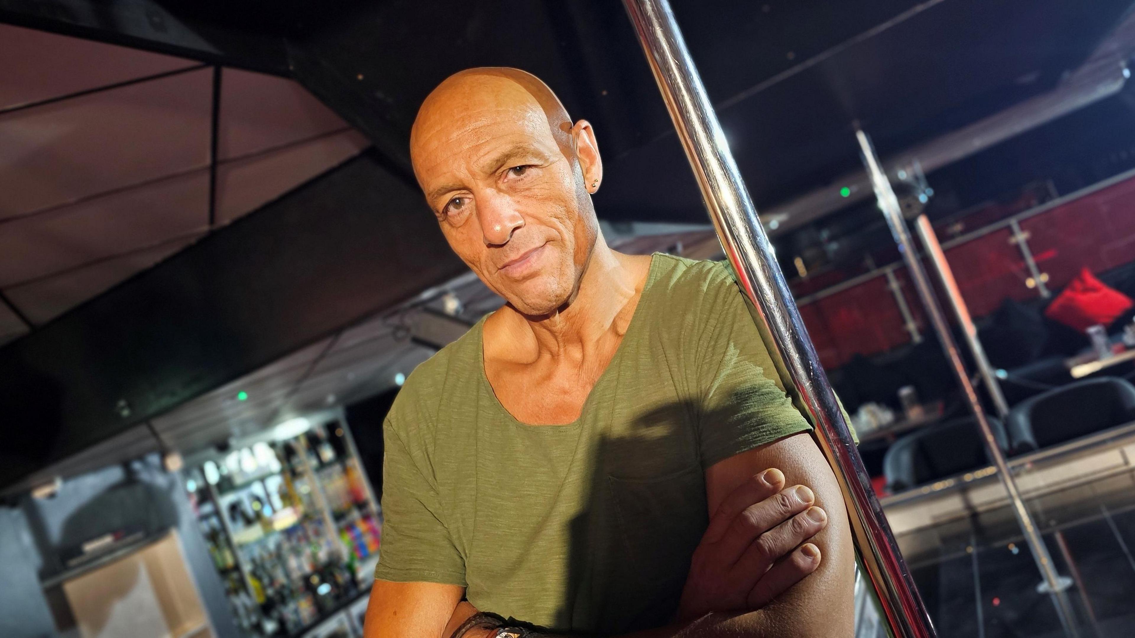 Daz Crawford posing in his club next to a dancing pole. His arms are folded and he's wearing a green t-shirt.