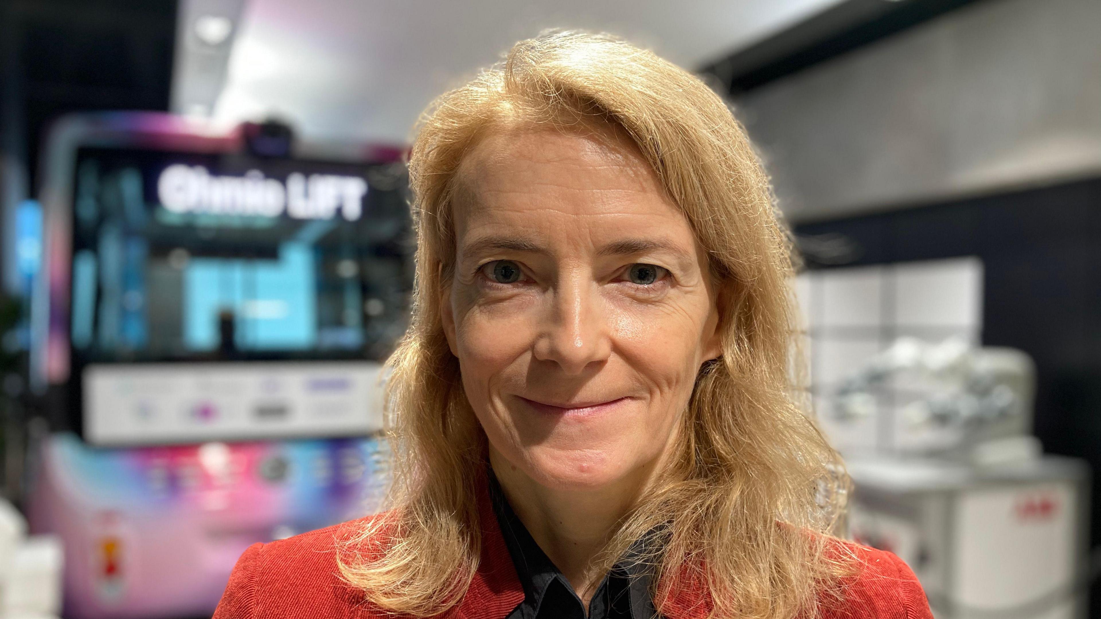 Liz Lloyd is smiling at the camera. She has blond air just past her shoulders and is wearing a red jacket with a black blouse underneath.
