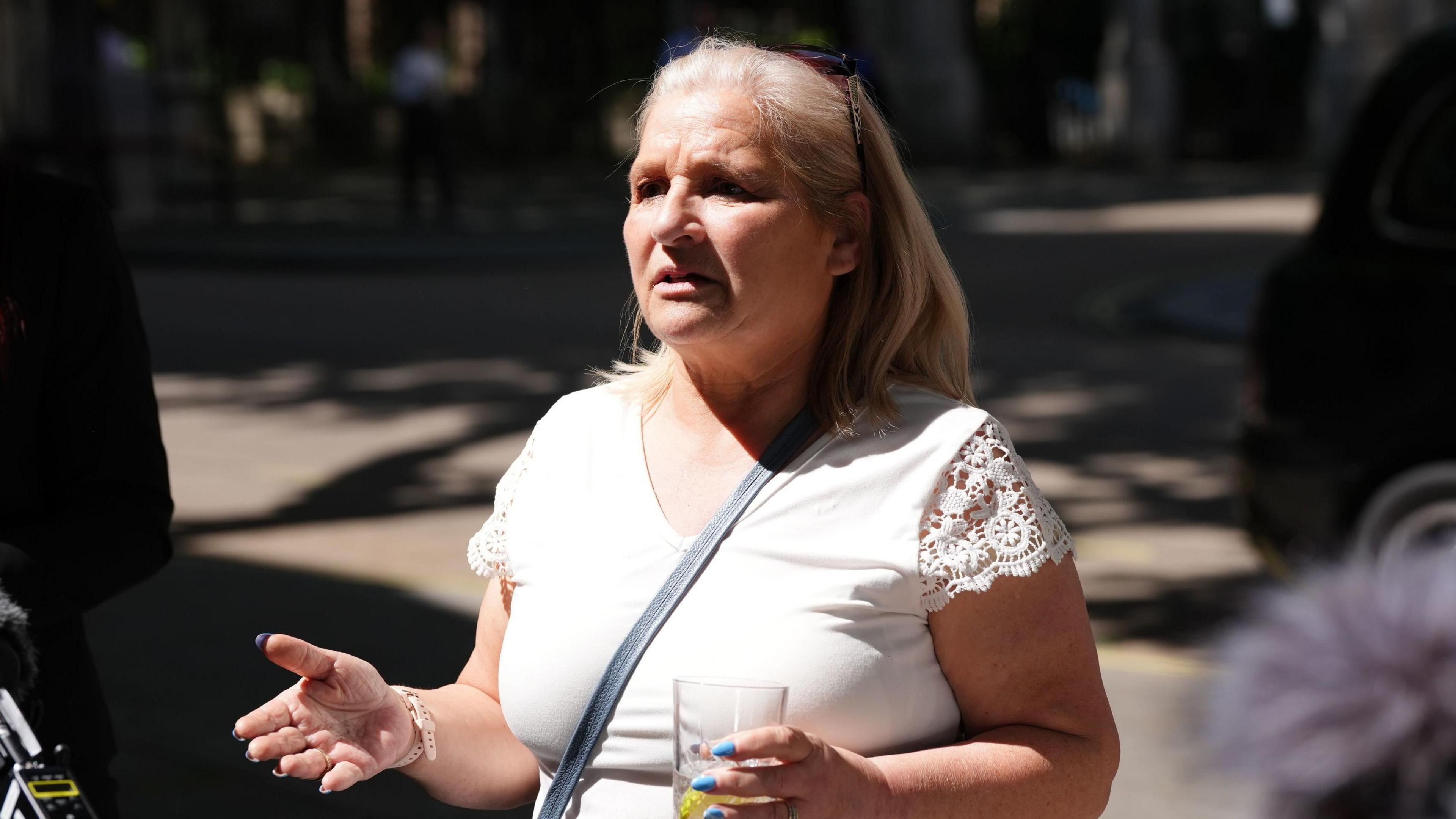 Kim Smith, who has blonde hair and is wearing a white top and has sunglasses perched on top of her head, speaks to reporters while holding a glass of water.