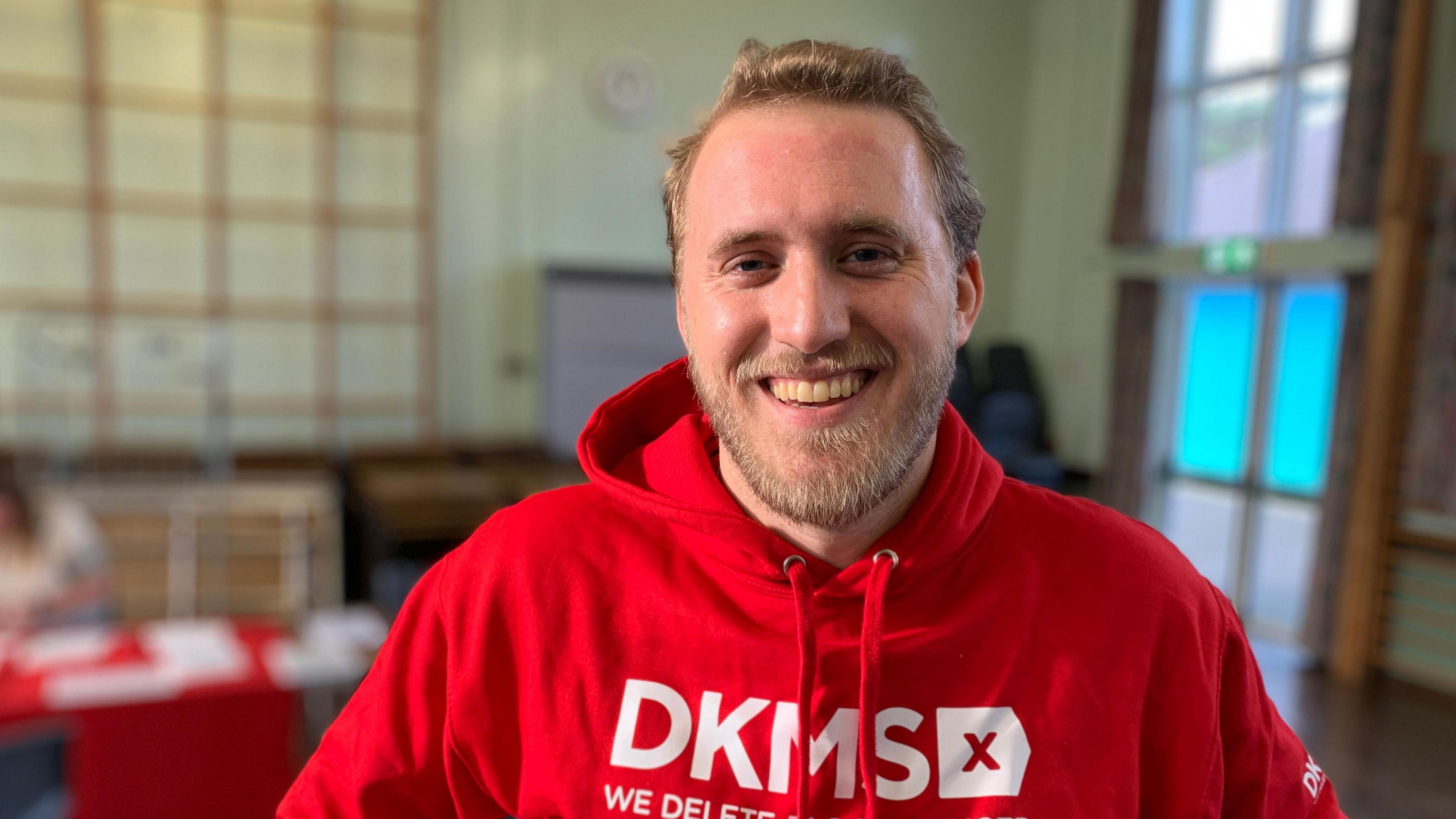 Michael Gallagher has short brown hair and a short brown beard. He is wearing a red hoody which has the letters DKMS emblazoned on it in white.