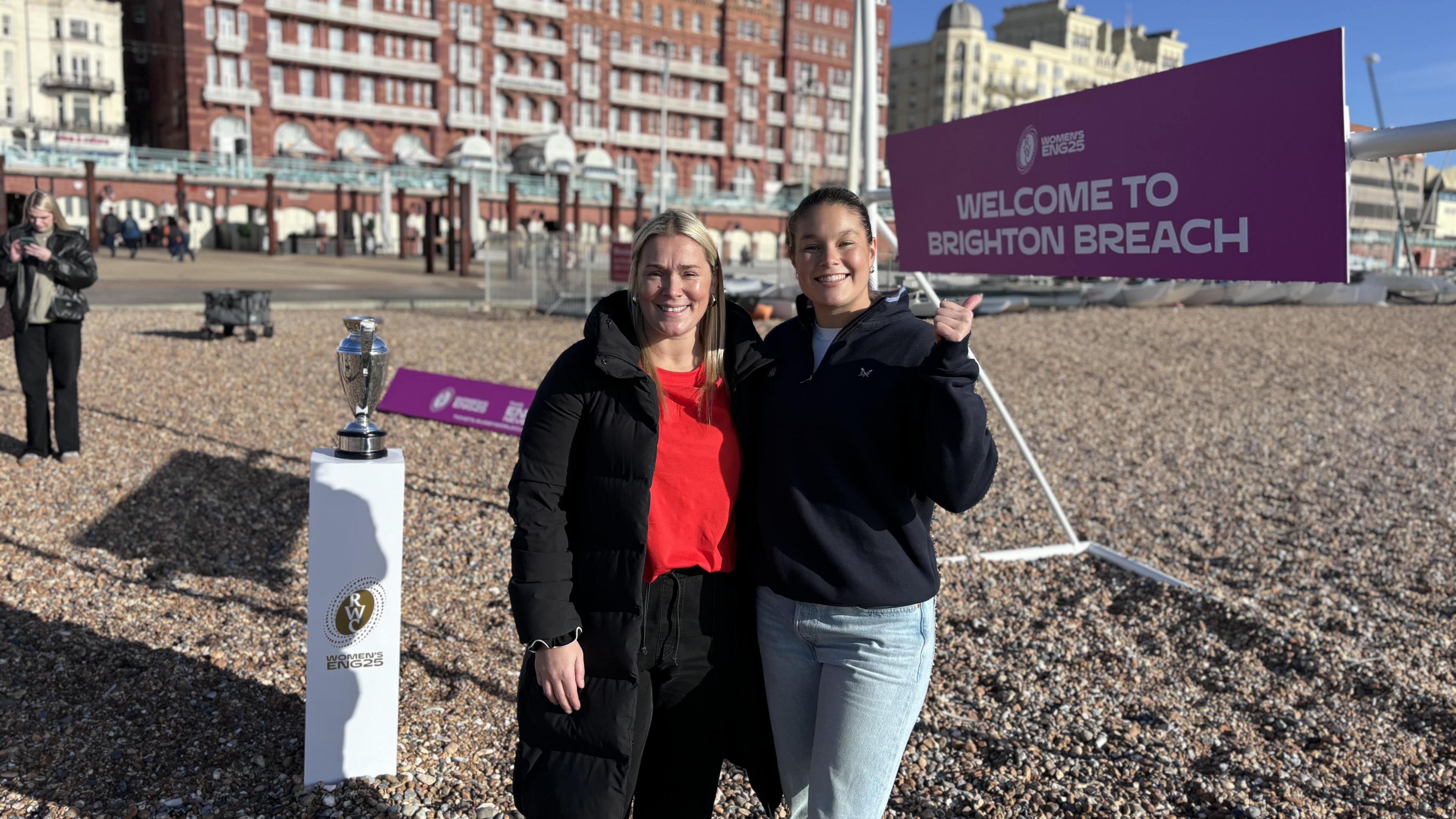 Jess Breach (right, a woman with light blue jeans, a dark blue jumper and her hair in a bun, smiling looking into the camera and holding a thumb up) and Rachael Burford (left, a woman with blonde hair and a long black coat with a red top on and smiling, looking into the camera) standing in front of a purple sign with white writing on saying Welcome To Brighton Breach on Brighton beach and a replica Woman' Rugby World Cup trophy trophy in the background