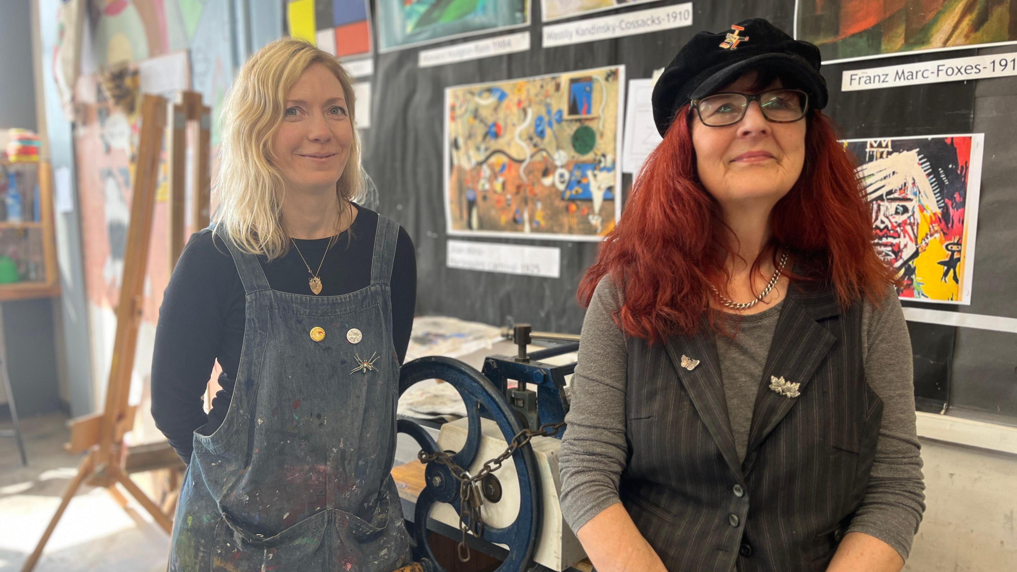 Two women stand in front of a manual printing press in an art room