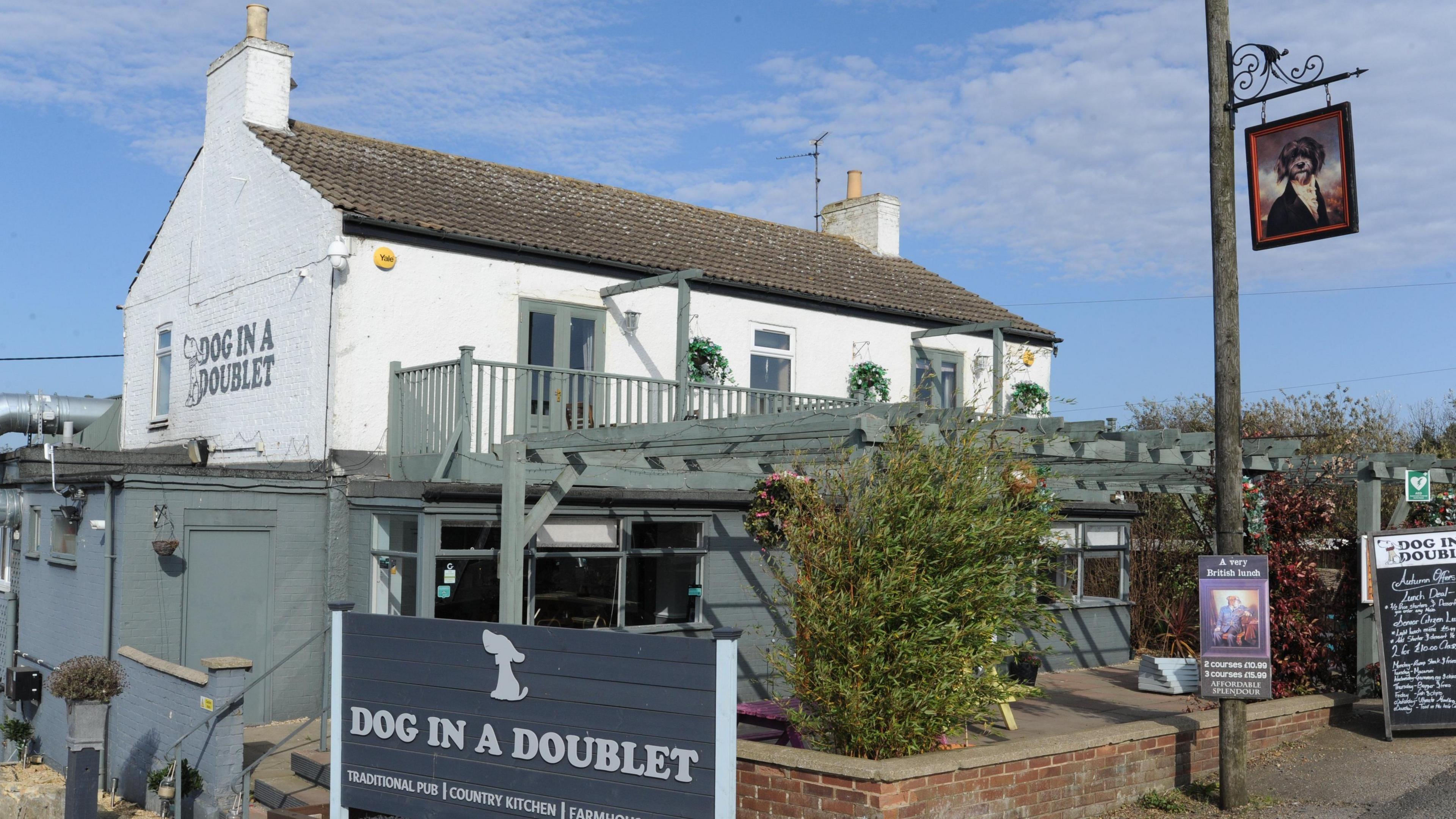 The exterior of the Dog in a Doublet pub. It is a white painted two-storey brick building, with green windows, balcony and pergola. Dog in a Doublet is written on the gable end of the pub, and in white letters on a grey sign in front of the building.