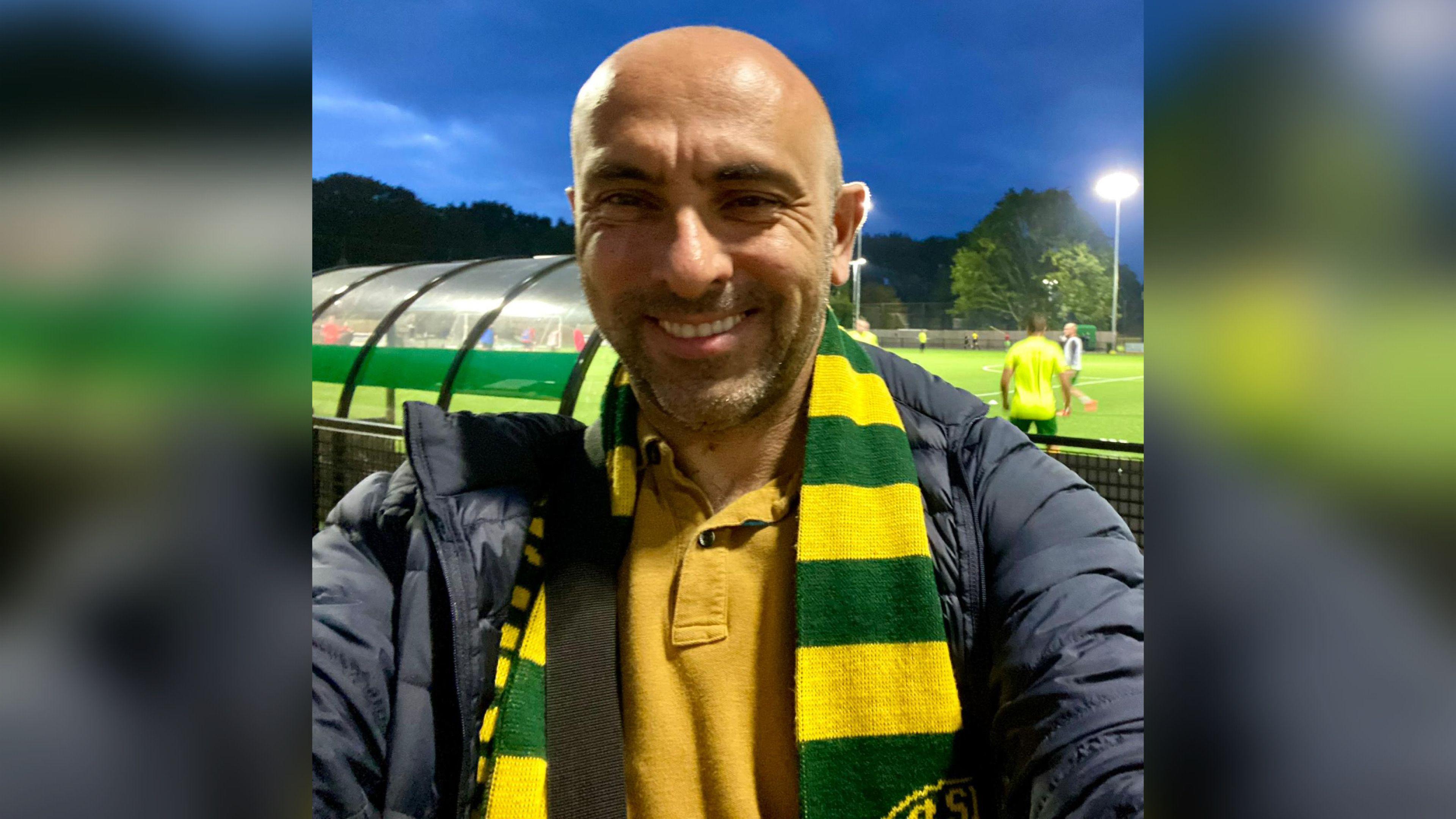A bald man with stubble smiling in a selfie. He is standing in front of a football pitch and has a yellow and green scarf round his neck.