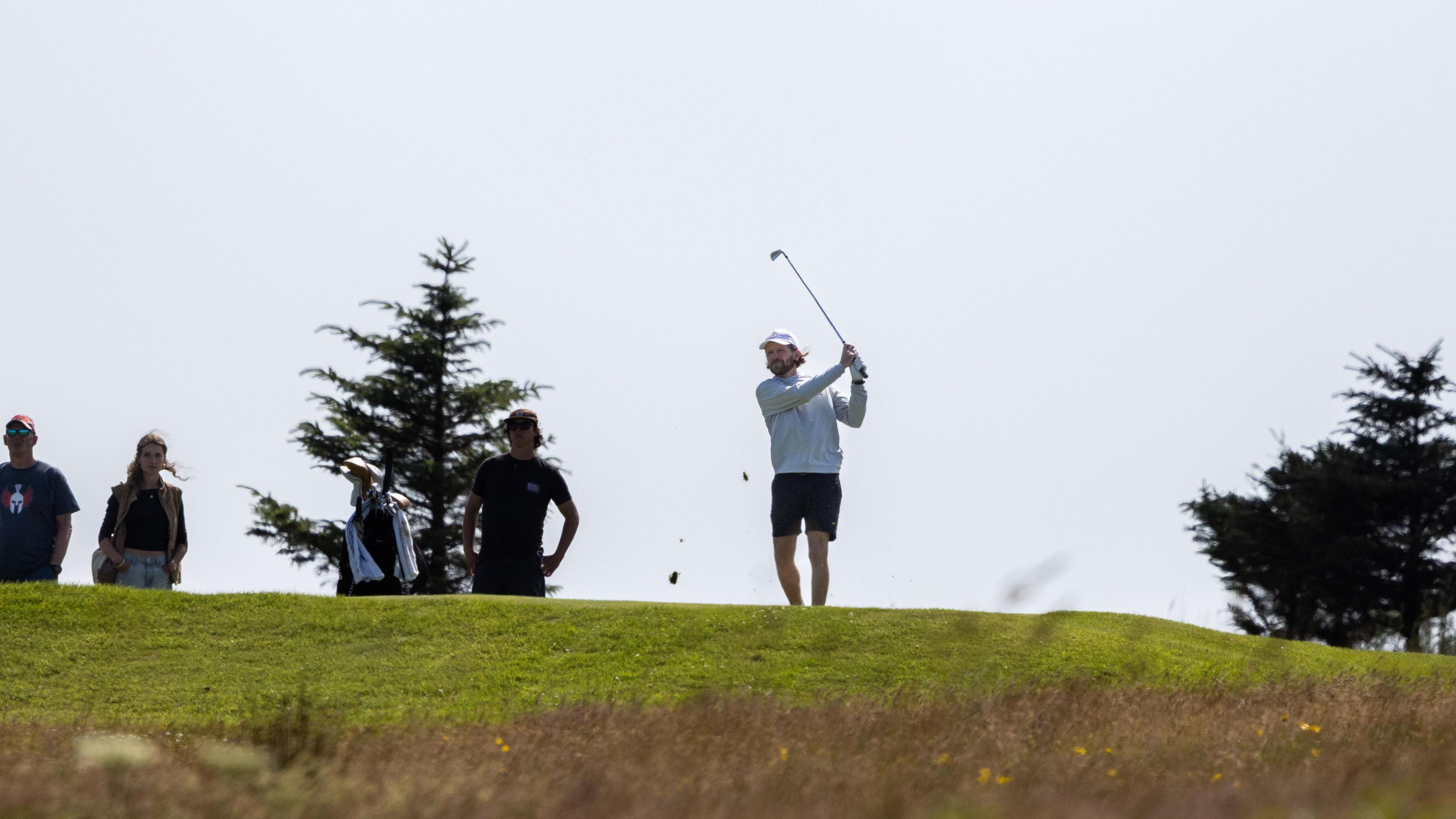 A man swings a golf club on a green course with large trees in the background. He wears dark shorts, a grey top and white cap. A man stands nearby, close to a bag full of clubs while another man and woman watch on.