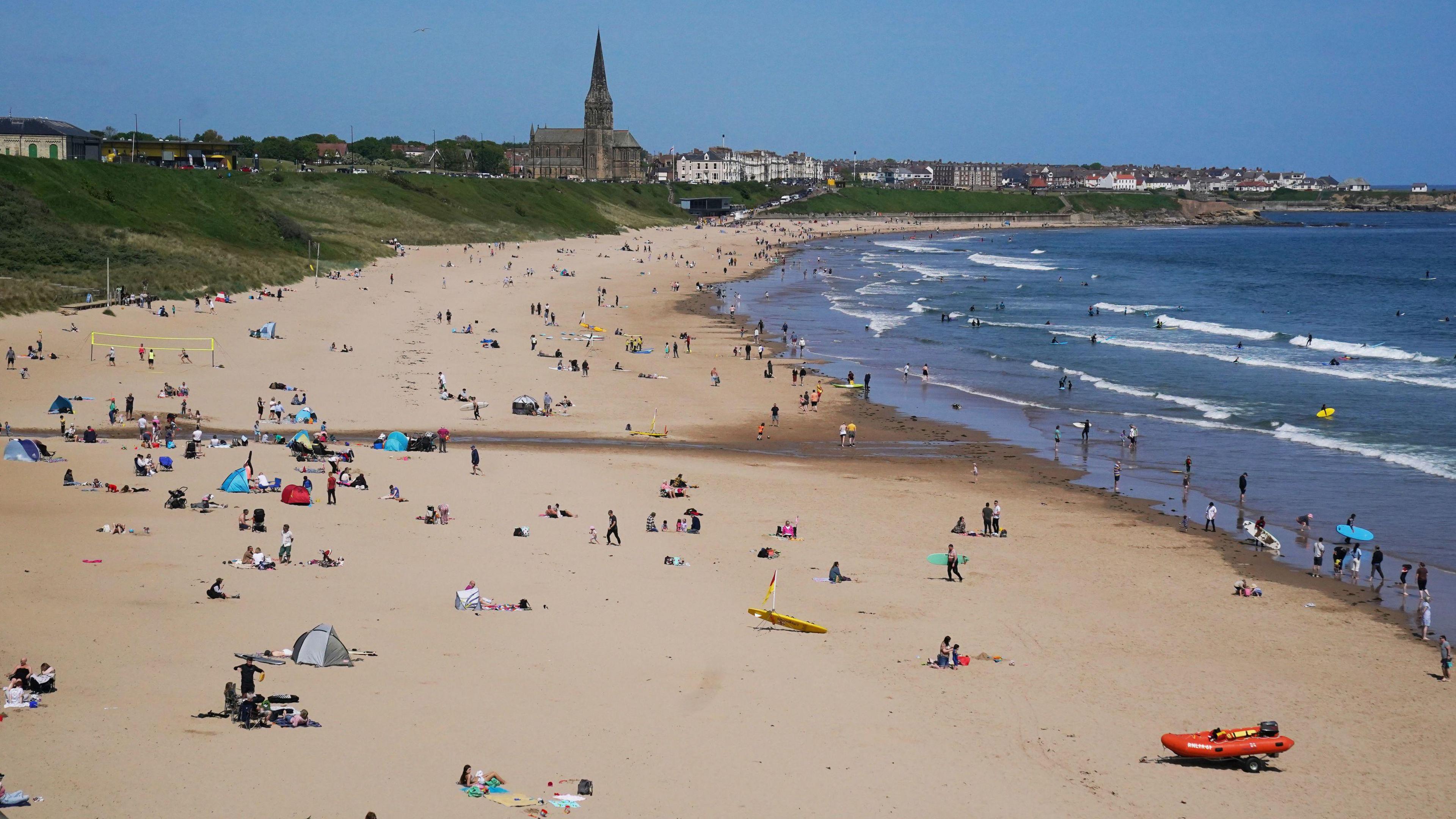 A busy beach filled with people relaxing, playing games on the yellow sand and going into the the blue sea. Red and yellow flags can be seen on the beach as well as rescue boards. A church can be seen on the street above the beach alongside houses.