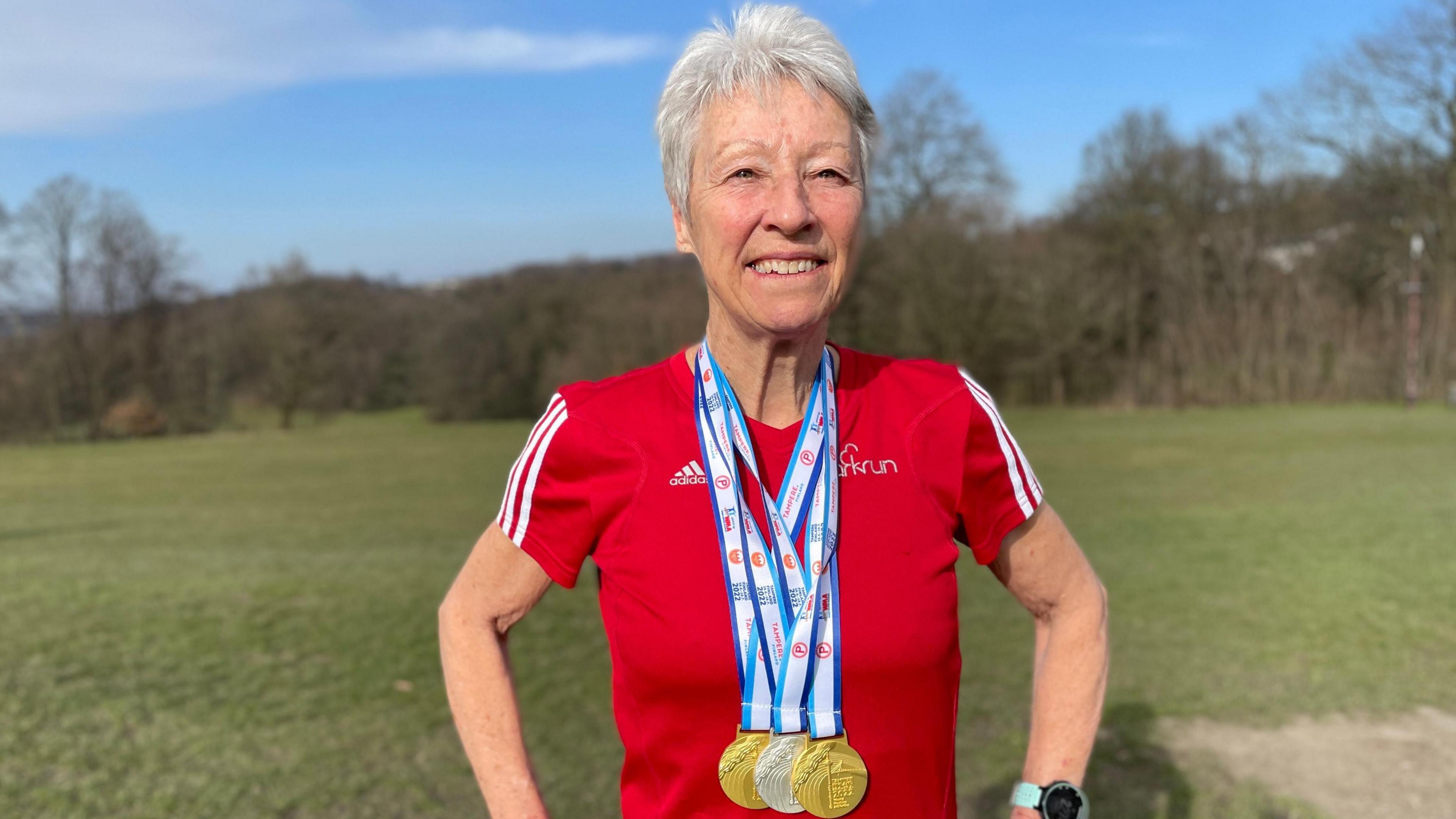 Dot Kesterton is wearing two gold and one silver medal round her neck from recent running races. She's smiling and has a red Parkrun t-shirt on.