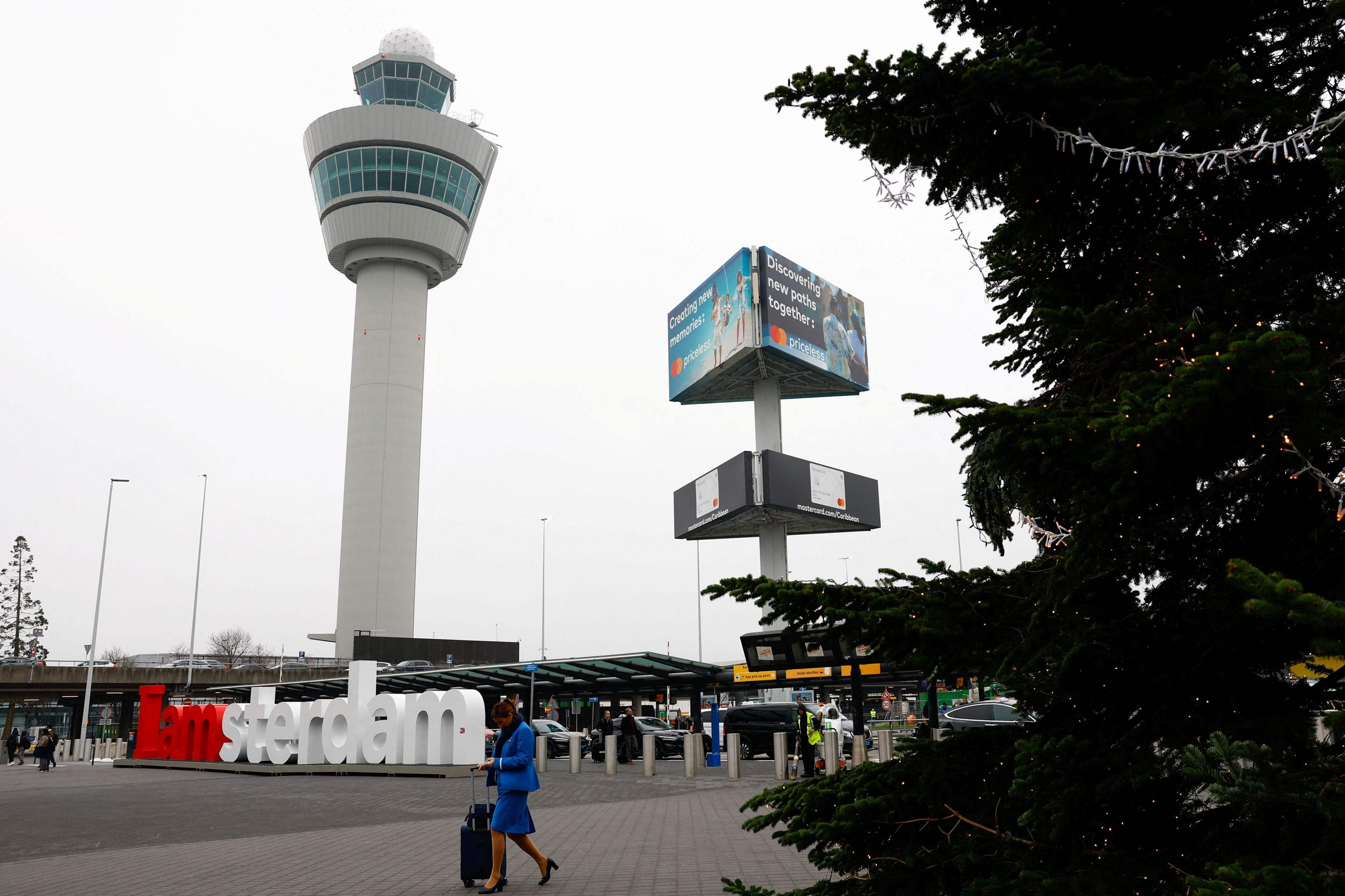 Schiphol airport in Amsterdam, with a large sign saying 'I am sterdam' and a woman in a blue suit standing next to an item of luggage, all in front of a white control tower