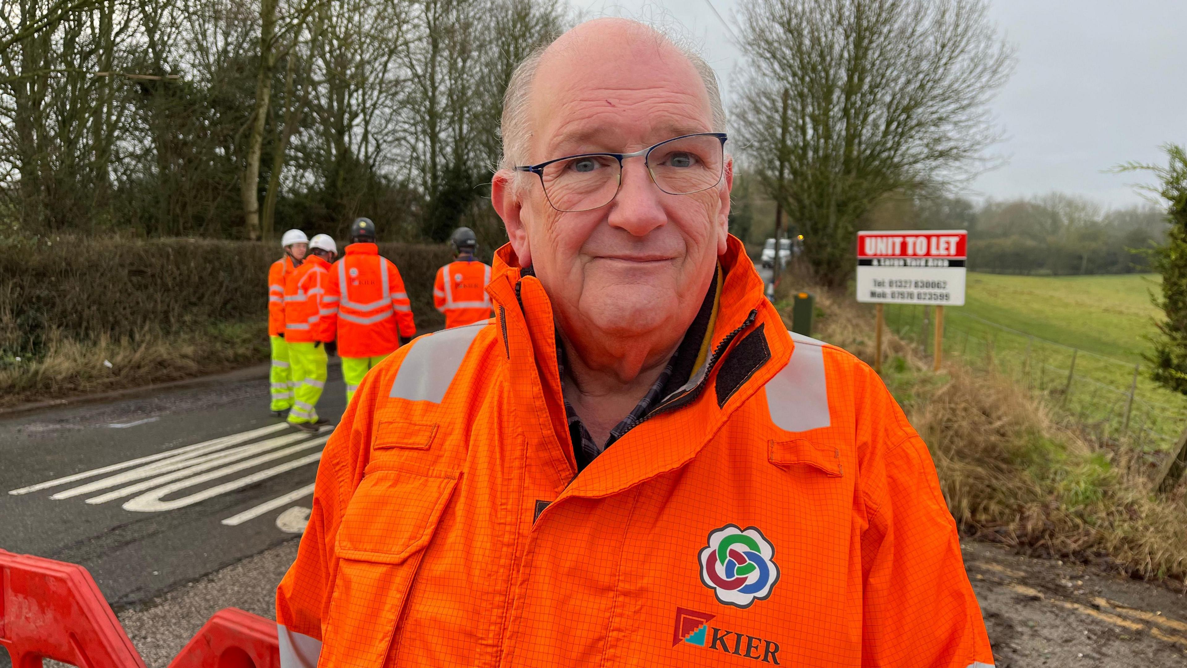 A man with glasses in a orange coat stands at the side of a road looking at the camera.