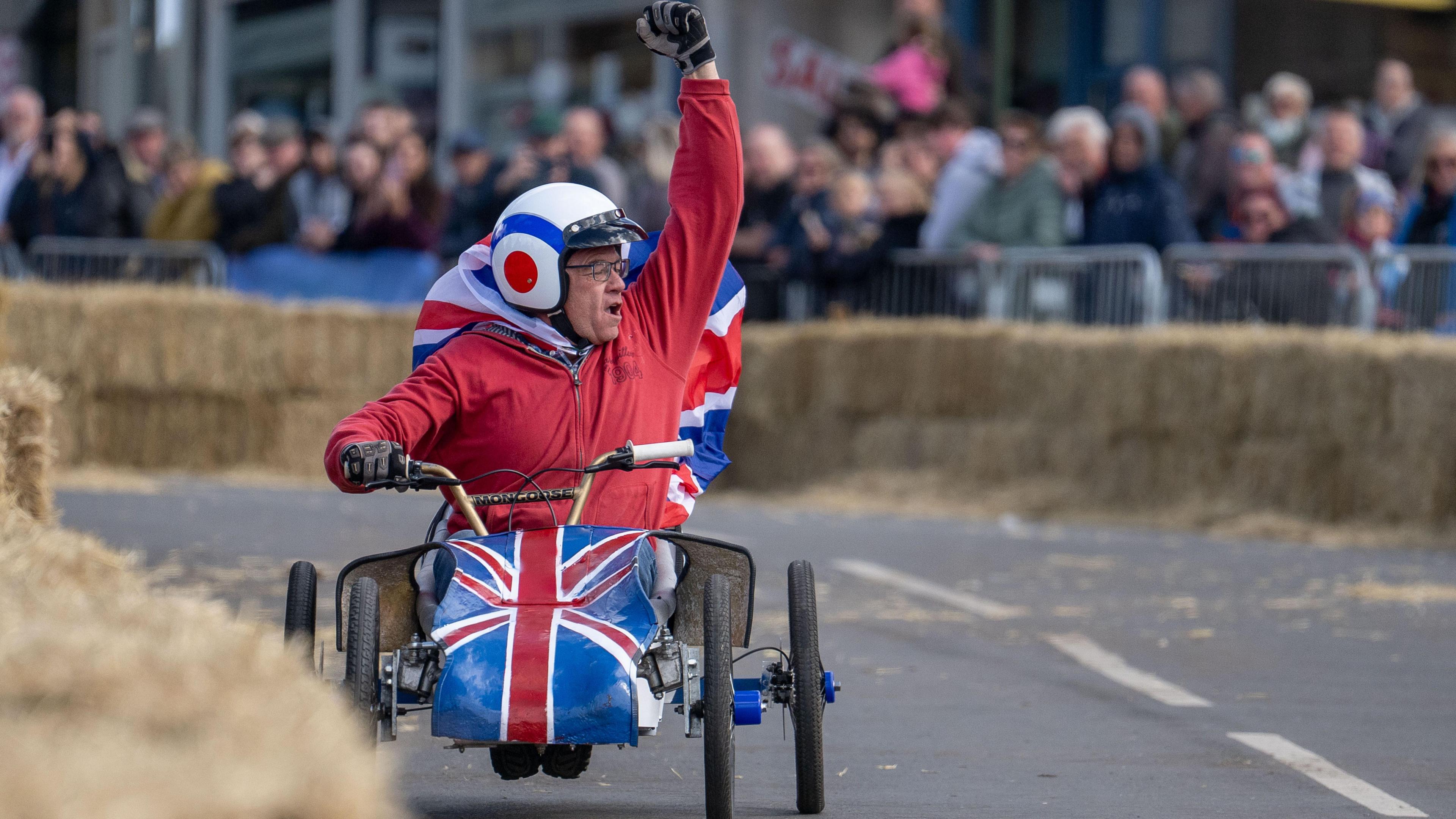 A driver in red overalls with a white helmet who is driving a kart which is painted with a union jack on the front of it. The driver jubilantly punches his fist into the air as spectators watch on.