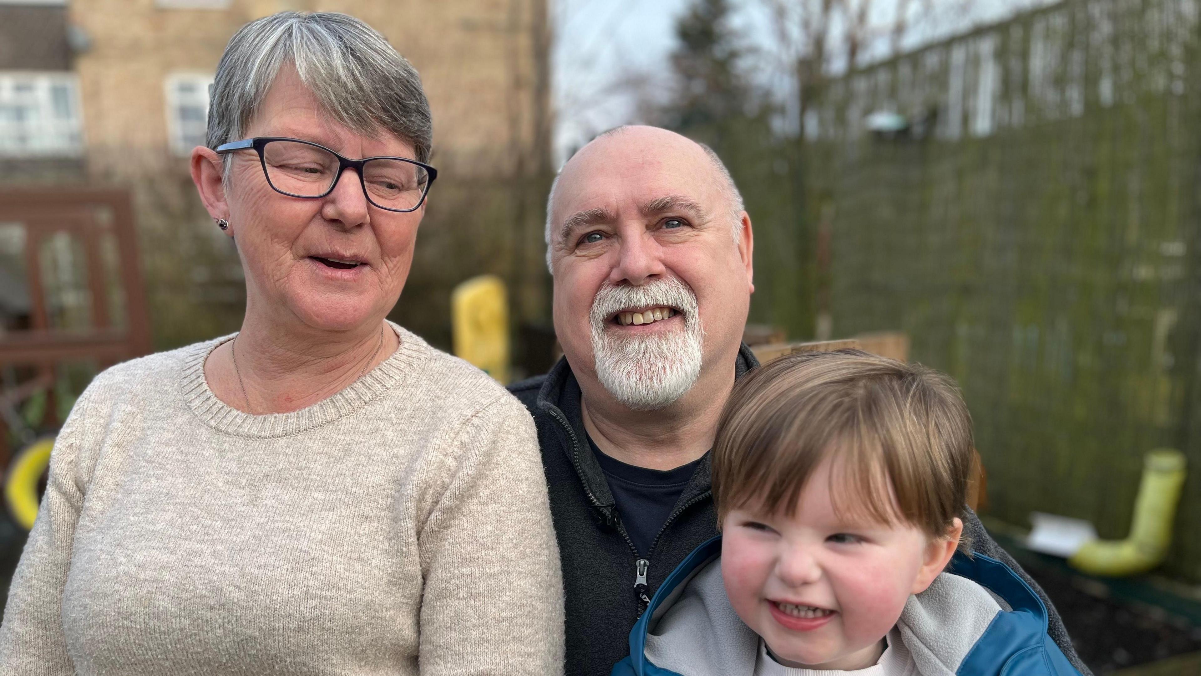 A family portrait of Tina, Chris and Hector. Chris is smiling directly at the camera, while Tina is smiling and looking down at Hector and Hector is looking to the left of the camera and smiling. Tina and Hector are sitting on Chris's lap, and they are photographed in a playground. 