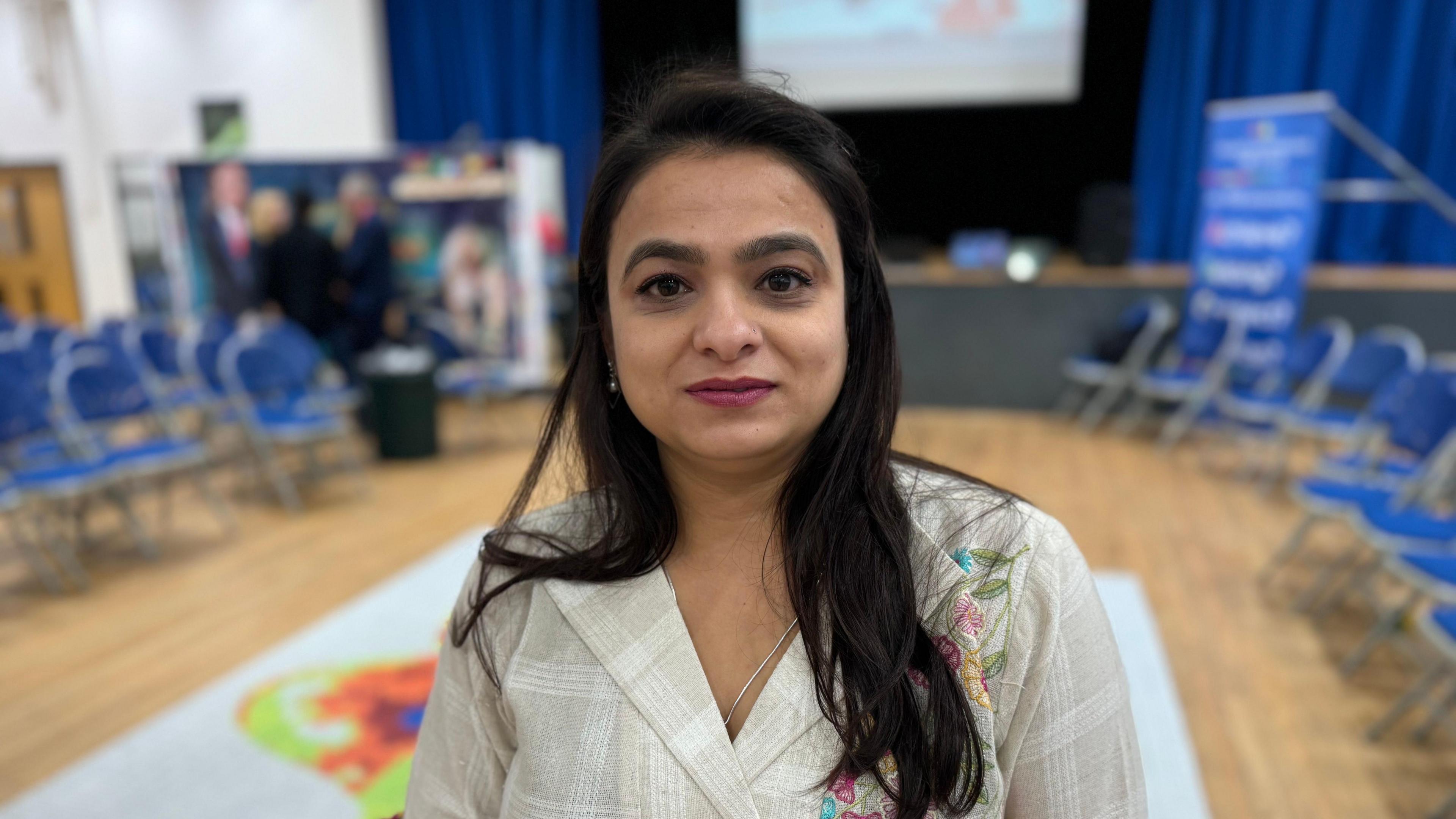 A woman looking straight at the camera. She is standing in a school hall and is wearing a white top with embroidered flowers.