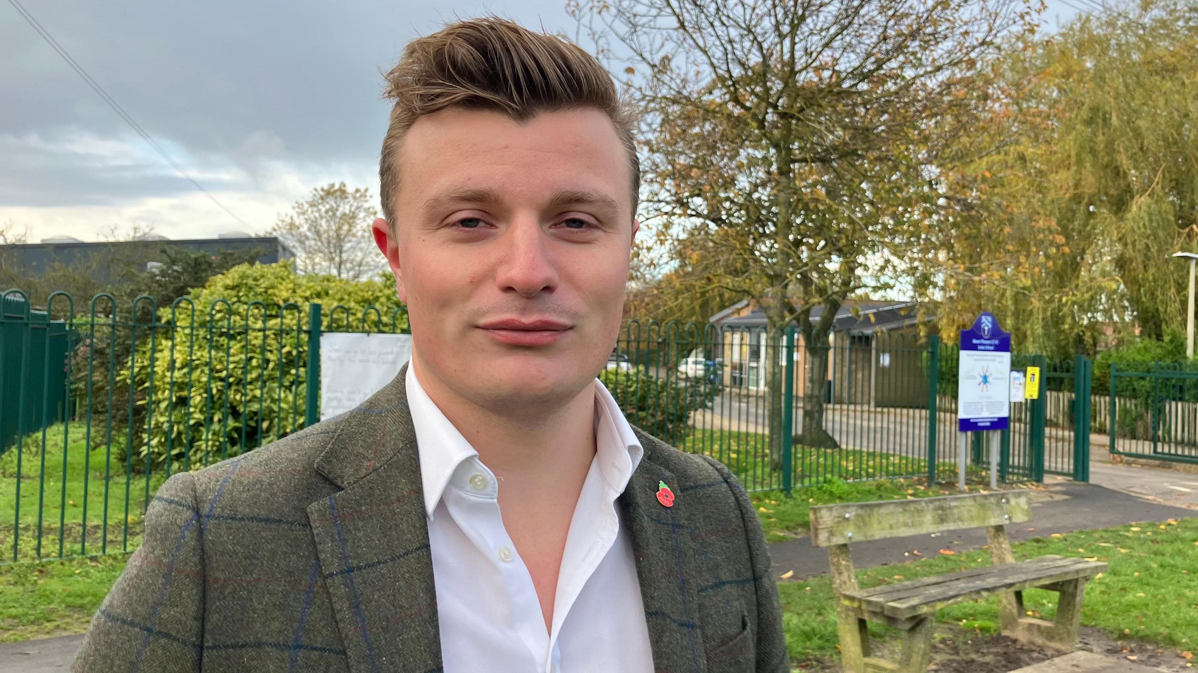 A man with a quiff in a suit stood outside a primary school looking down the barrel of the camera