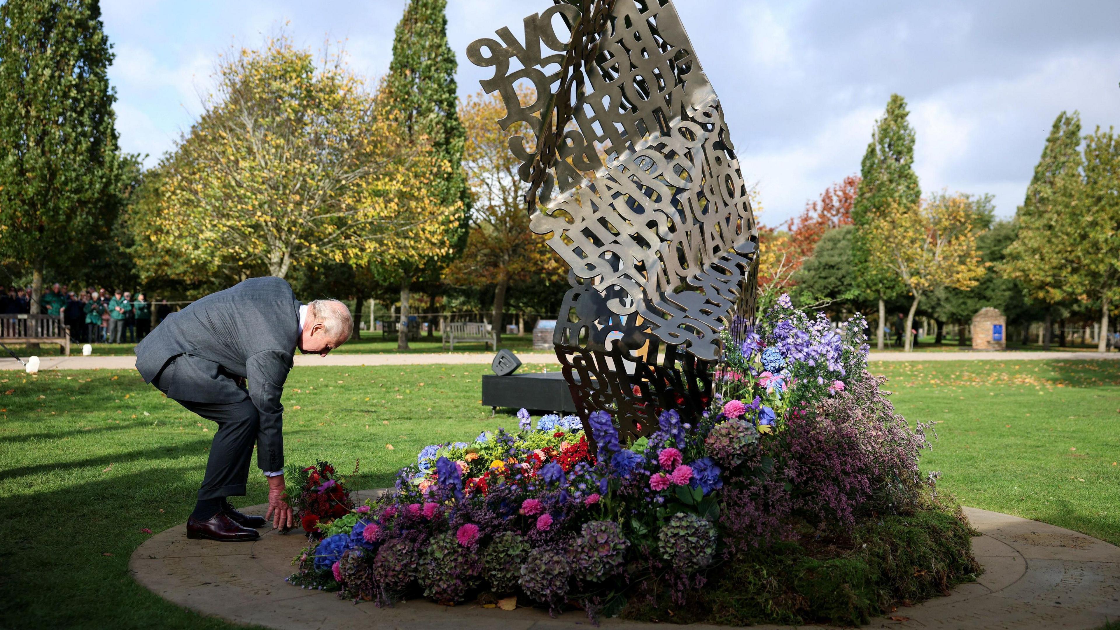 A wide shot of King Charles III laying flowers in front of the memorial. The sculpture is a collection of different words.