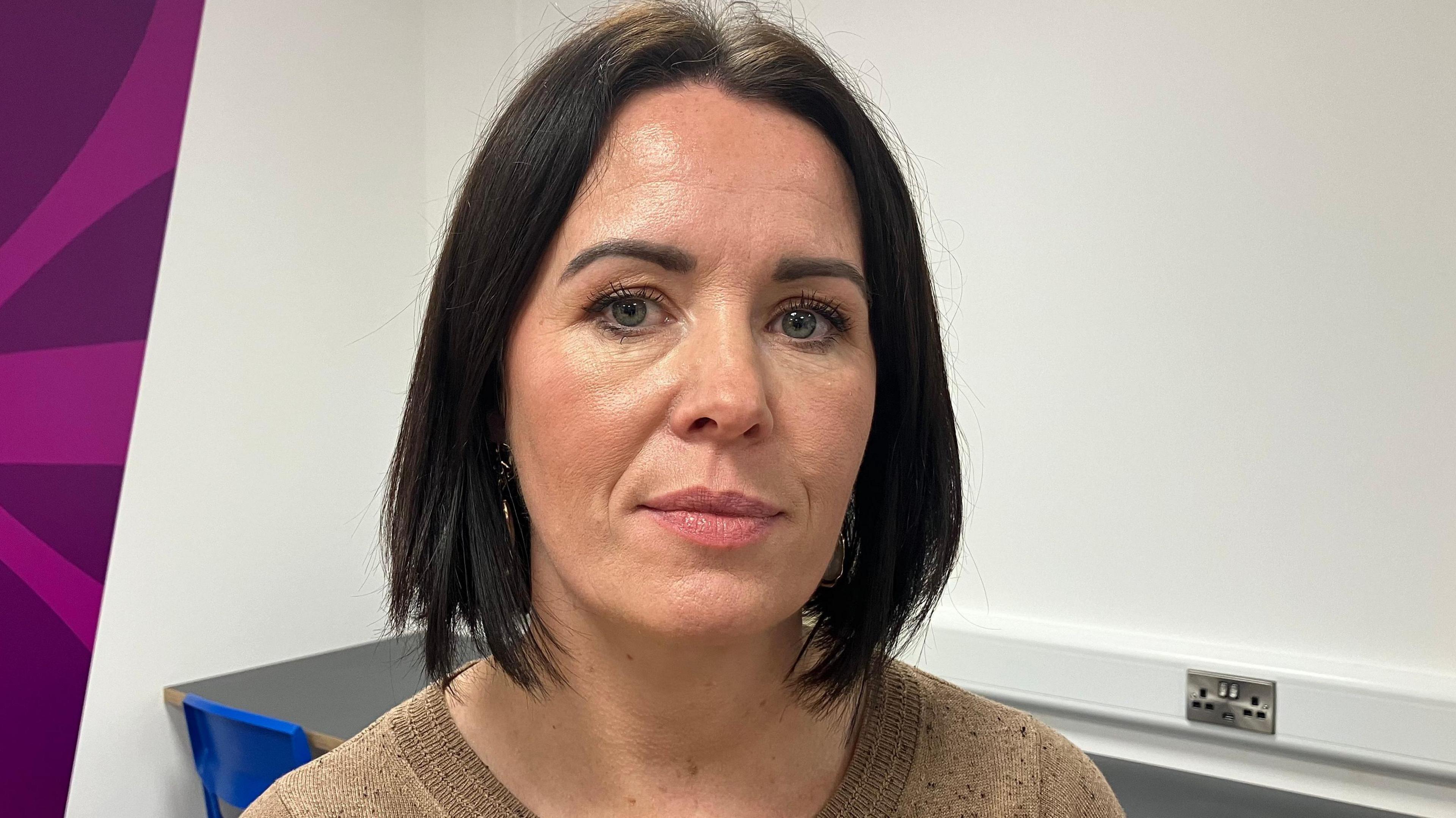 A woman, with black shoulder length hair, looking at the camera. She is wearing a cream coloured top. There is a white wall behind her. 