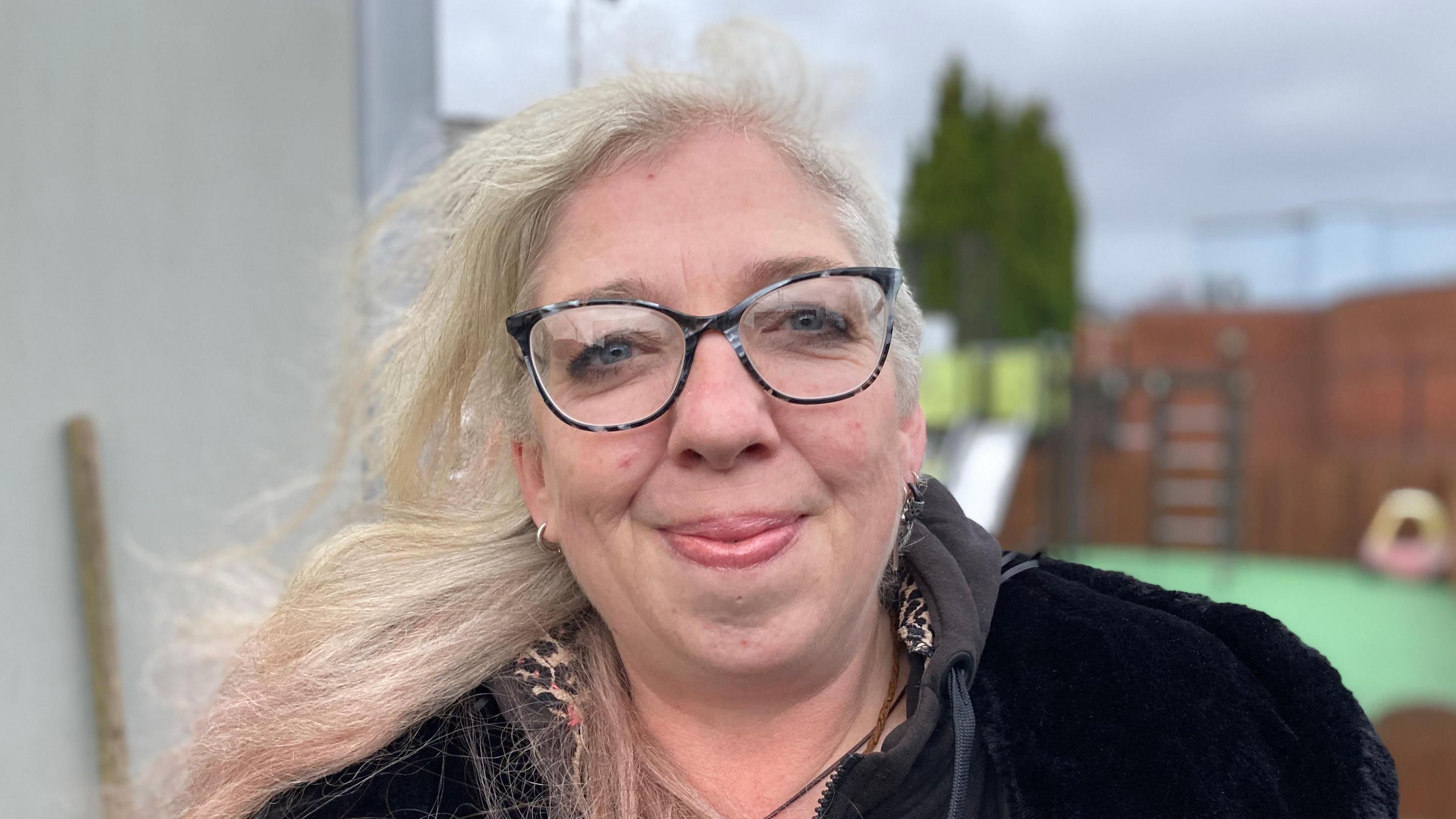 Emily Gallagher has long blonde hair and wears black glasses. She is smiling in a close-up shot as she wears a black coat.