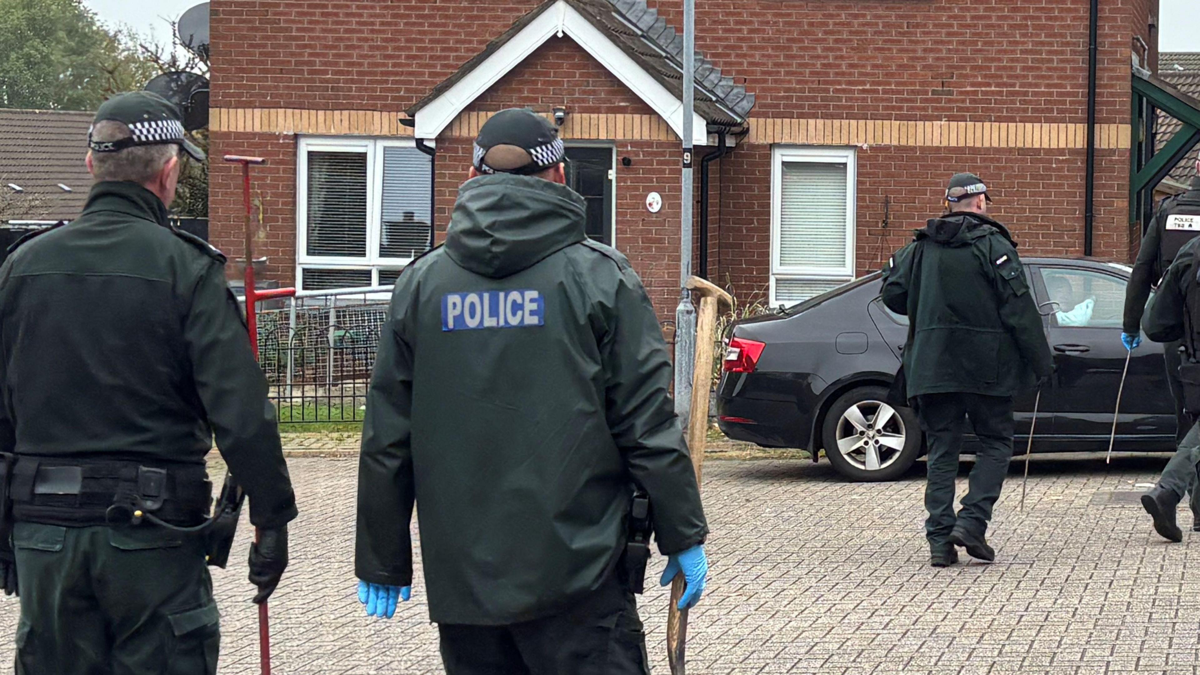 Five police officers are walking towards red brick houses. The house are two stories high with grey roofs. The officers are all wearing green and carrying items. There are two cars parked, one black and one silver. In the distance there are more house roofs visible.