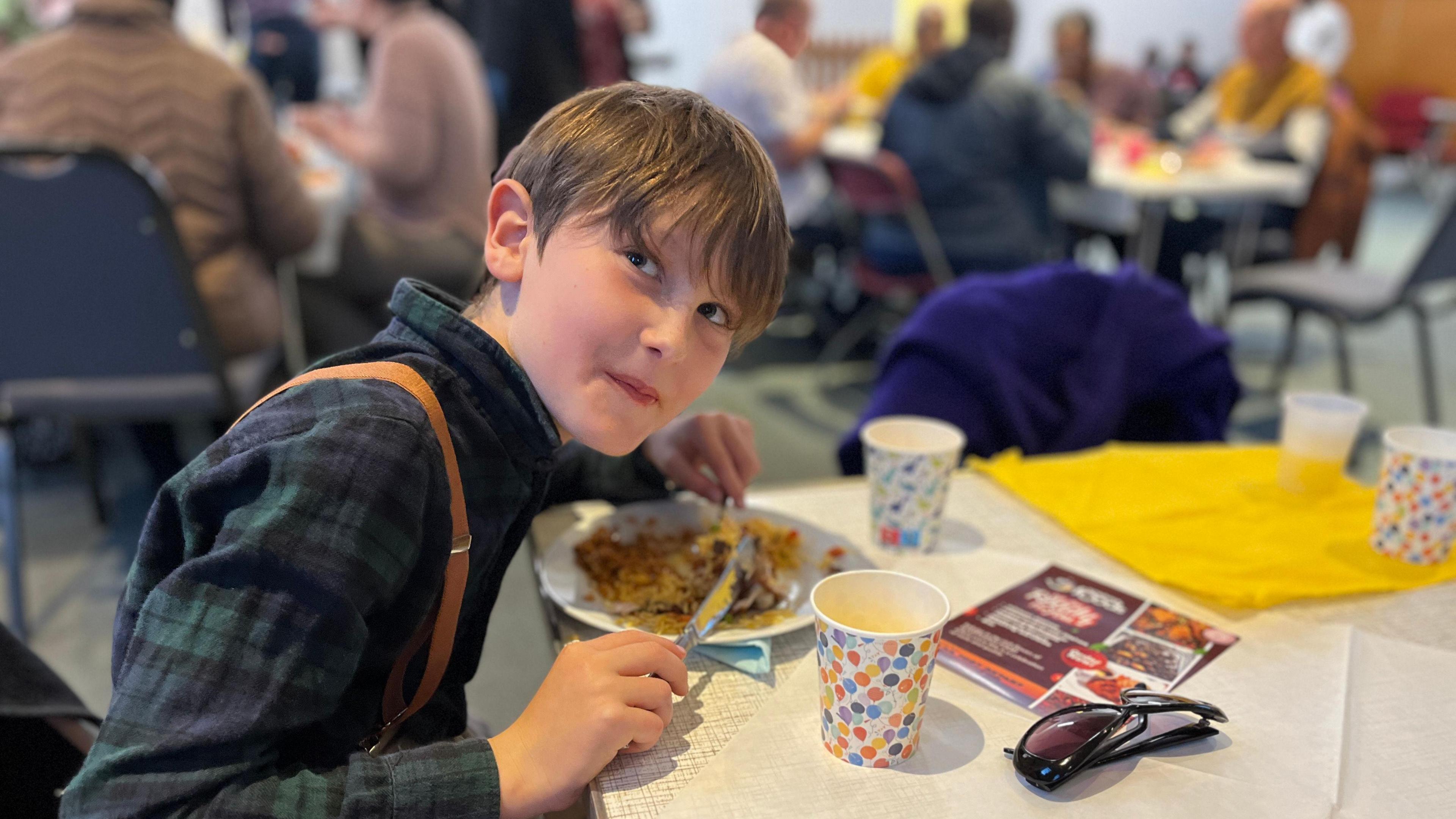 A 10-year-old boy sits at a table happily eating a plate of food.