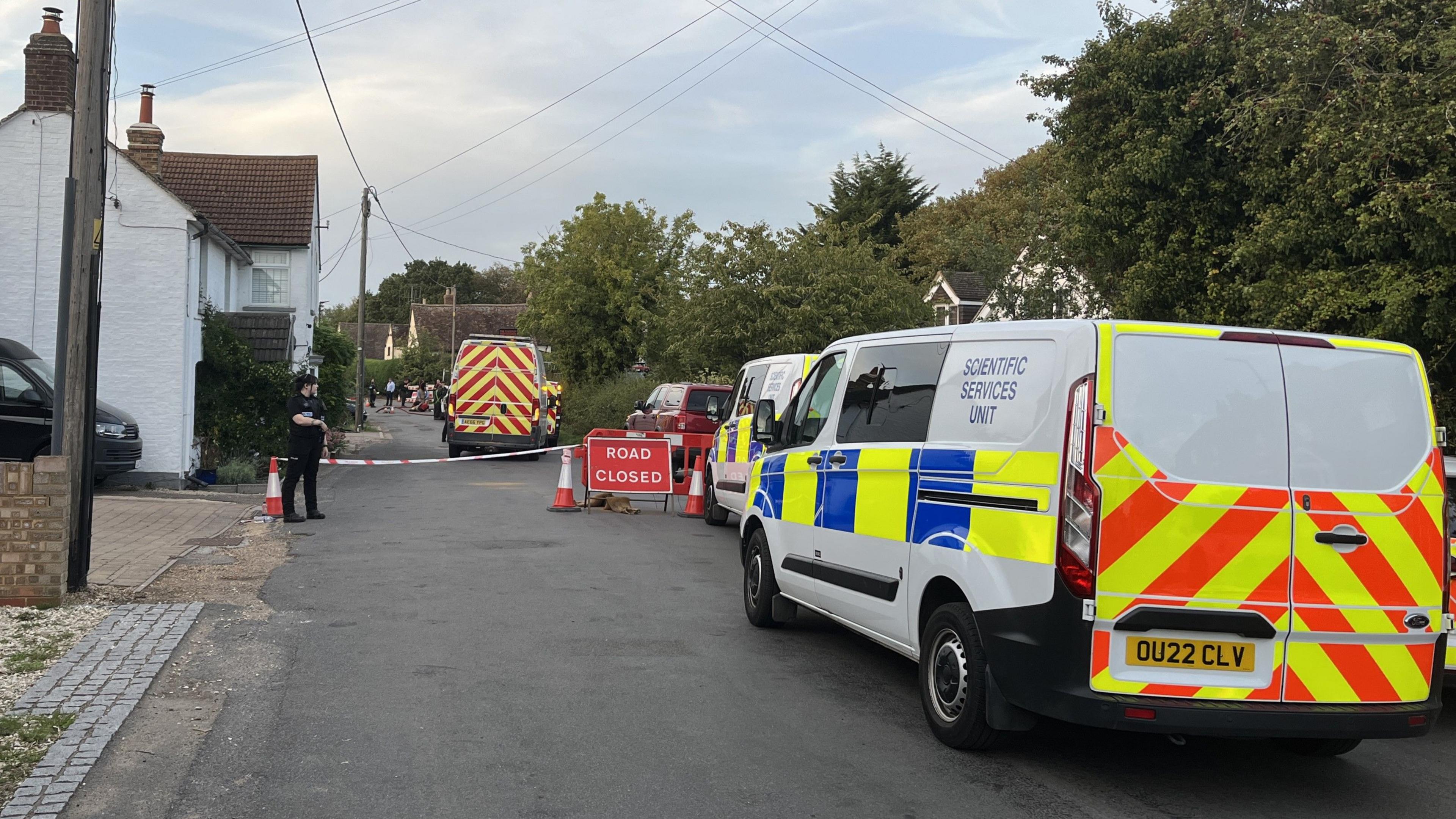 A police forensics van is parked on a road with another in front of it. There is a police cordon in the distance which is manned by a uniformed police officer. Other emergency service vehicles can be seen within the cordon. 