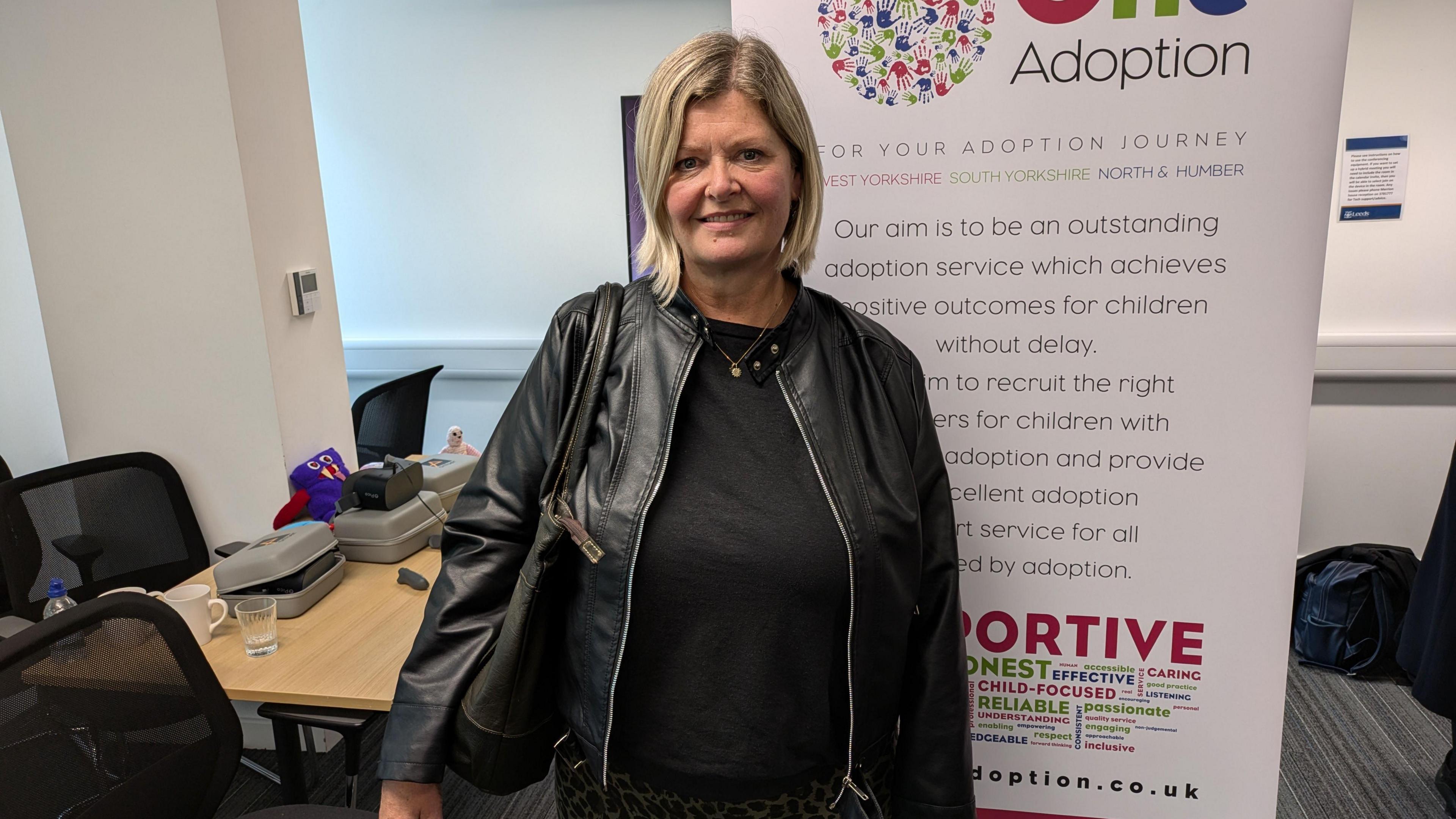 A woman in her fifties with blonde hair and a black leather coat worn over the top of a black top. She is stood partially in front of an advertising board for One Adoption, which has large writing on explaining what the agency is. A desk with various items is over her right shoulder.