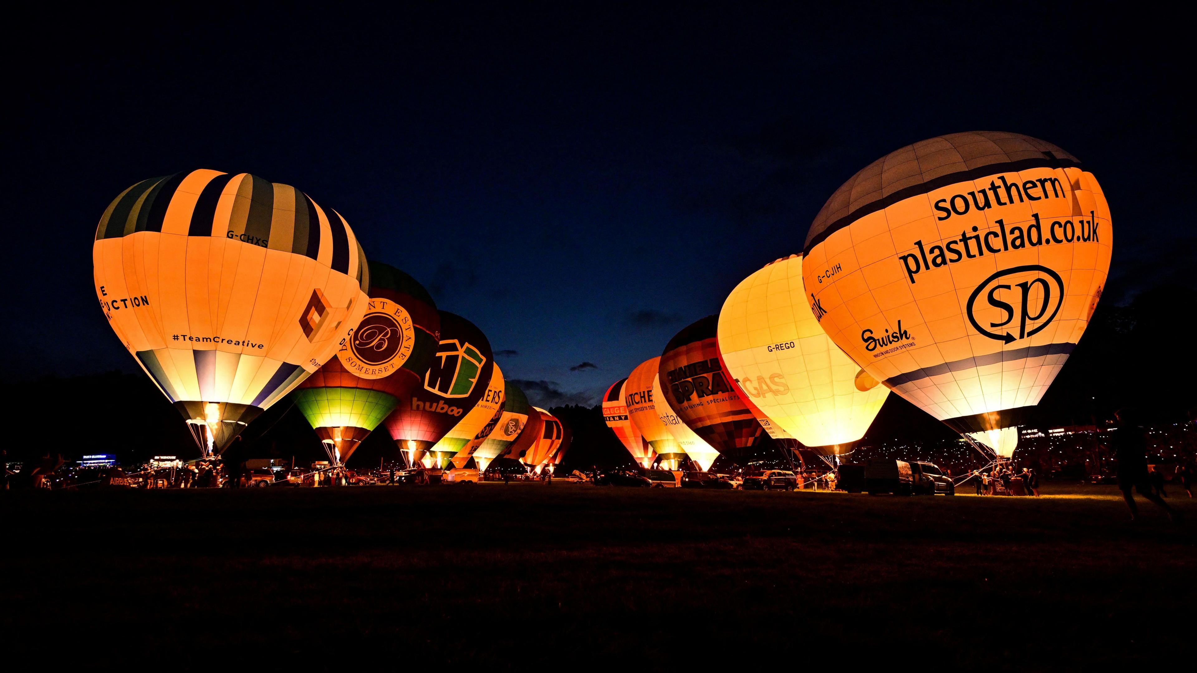 Two lines of hot air balloons tethered to the ground. They are all lit up from their burners. The image is taken at night time.