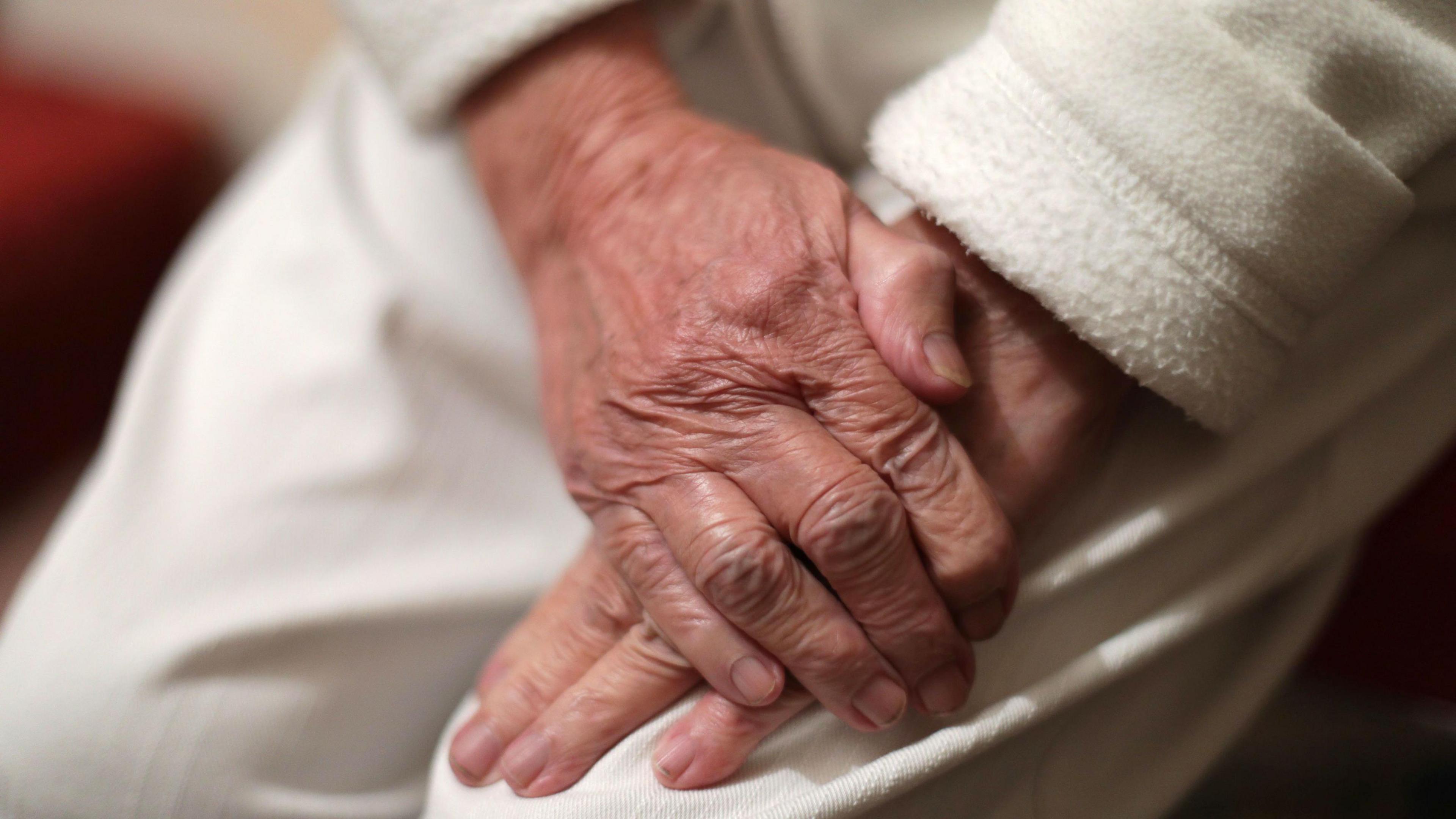 Close up on: An old person in a dressing gown with papery skin clenches one hand with the other.