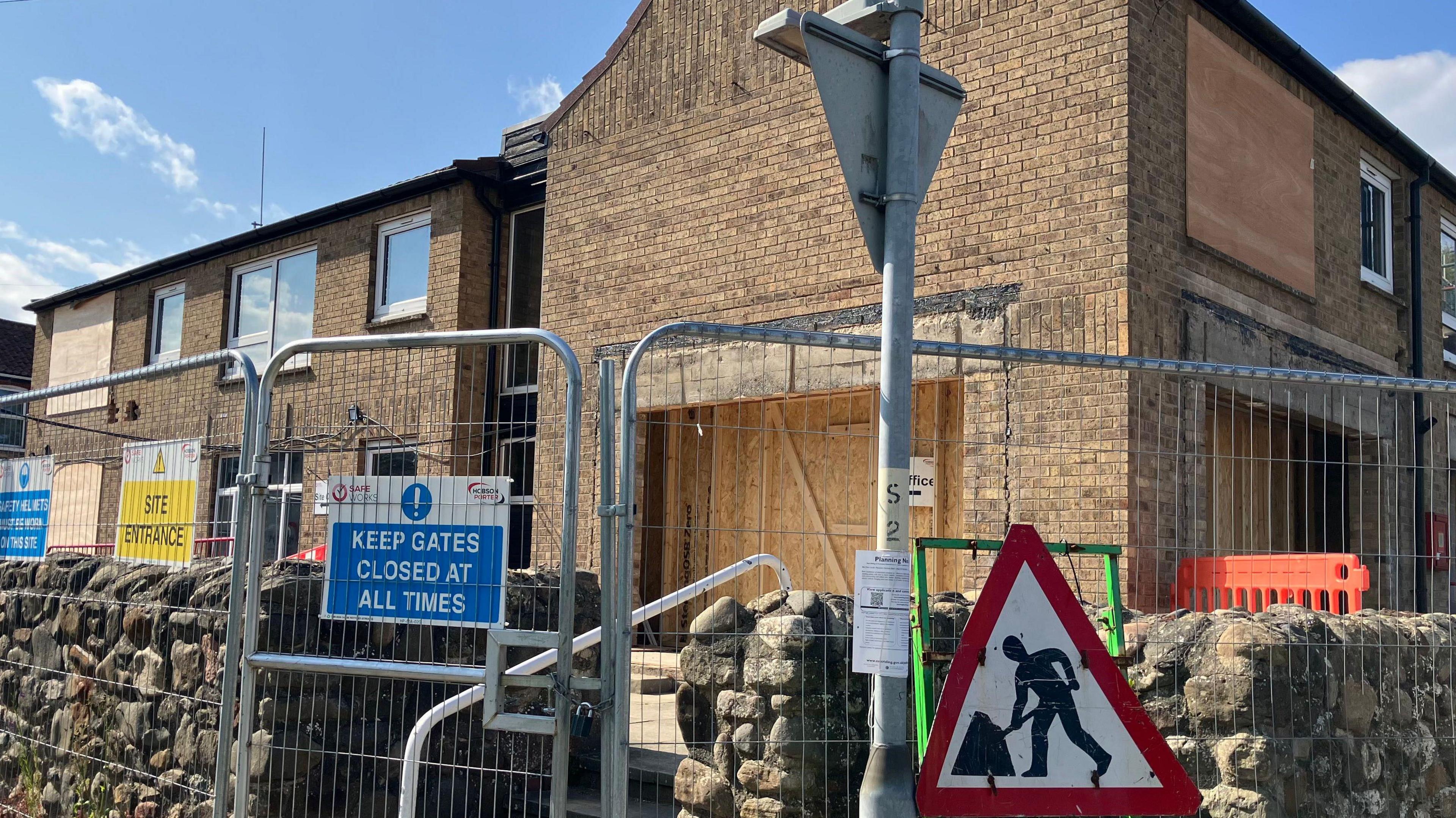 View of a boarded up two-storey building, There is a metal fence surrounding the site and a "men at work" road sign.