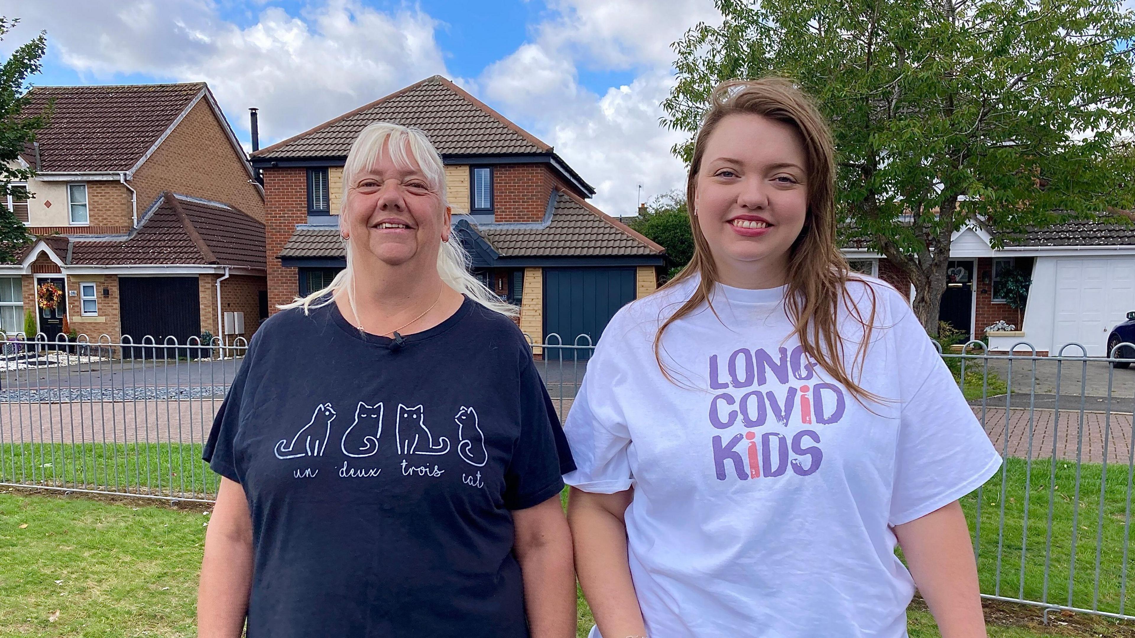 Sue and Anita Widdowson stand in a park and smile at the camera. Houses are in the background. Sue, who has white/blonde hair and a fringe, wears a dark t-shirt with the outline of four cats in white. The words "un deux trois cat" are written underneath the cats. Anita, who has long dark hair, is wearing a white t-shirt with the words "long covid kids" written in mostly purple text.