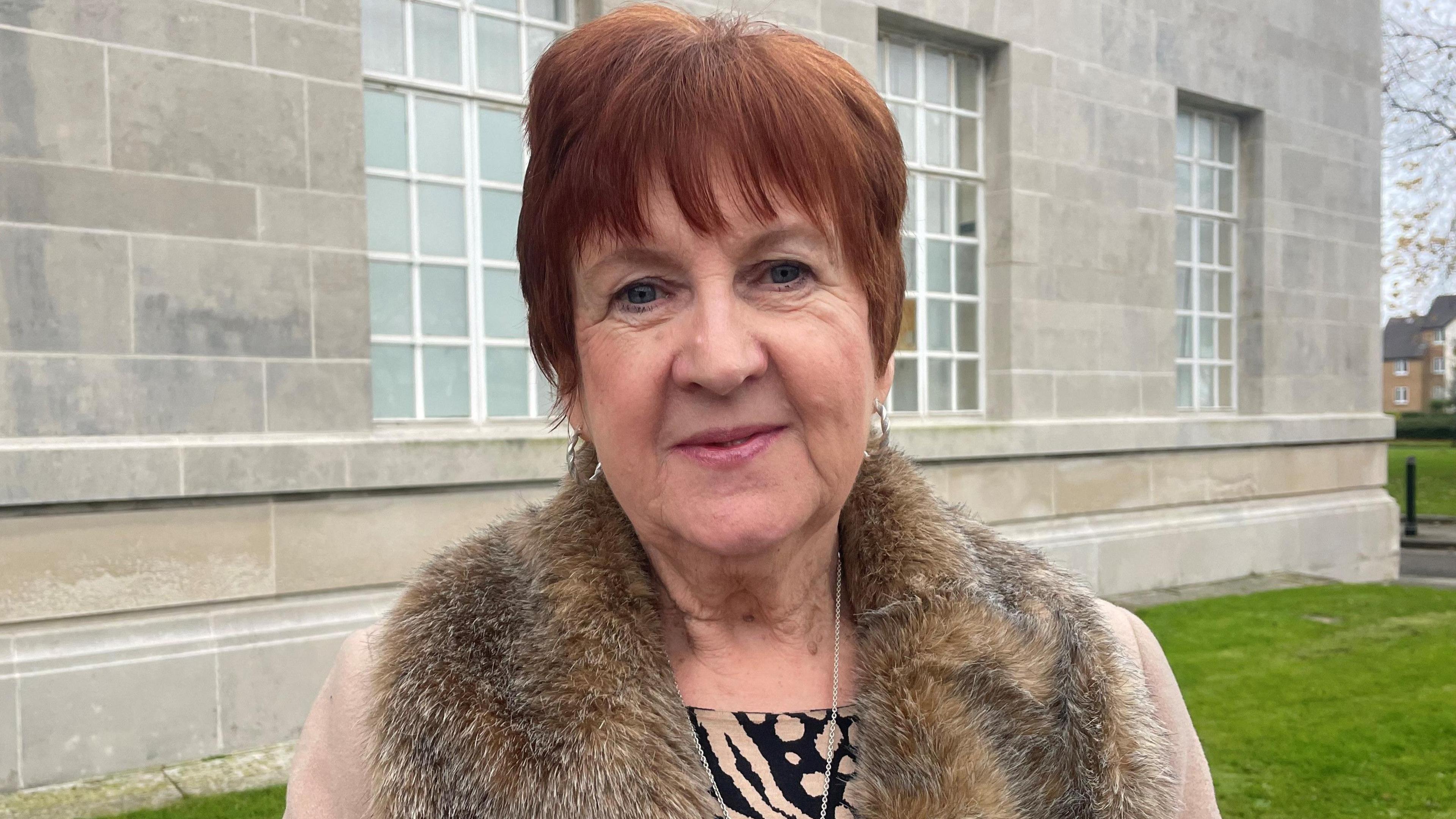 A woman with cropped red hair, wearing a fur-looking jacket. She is standing in front of a grey building