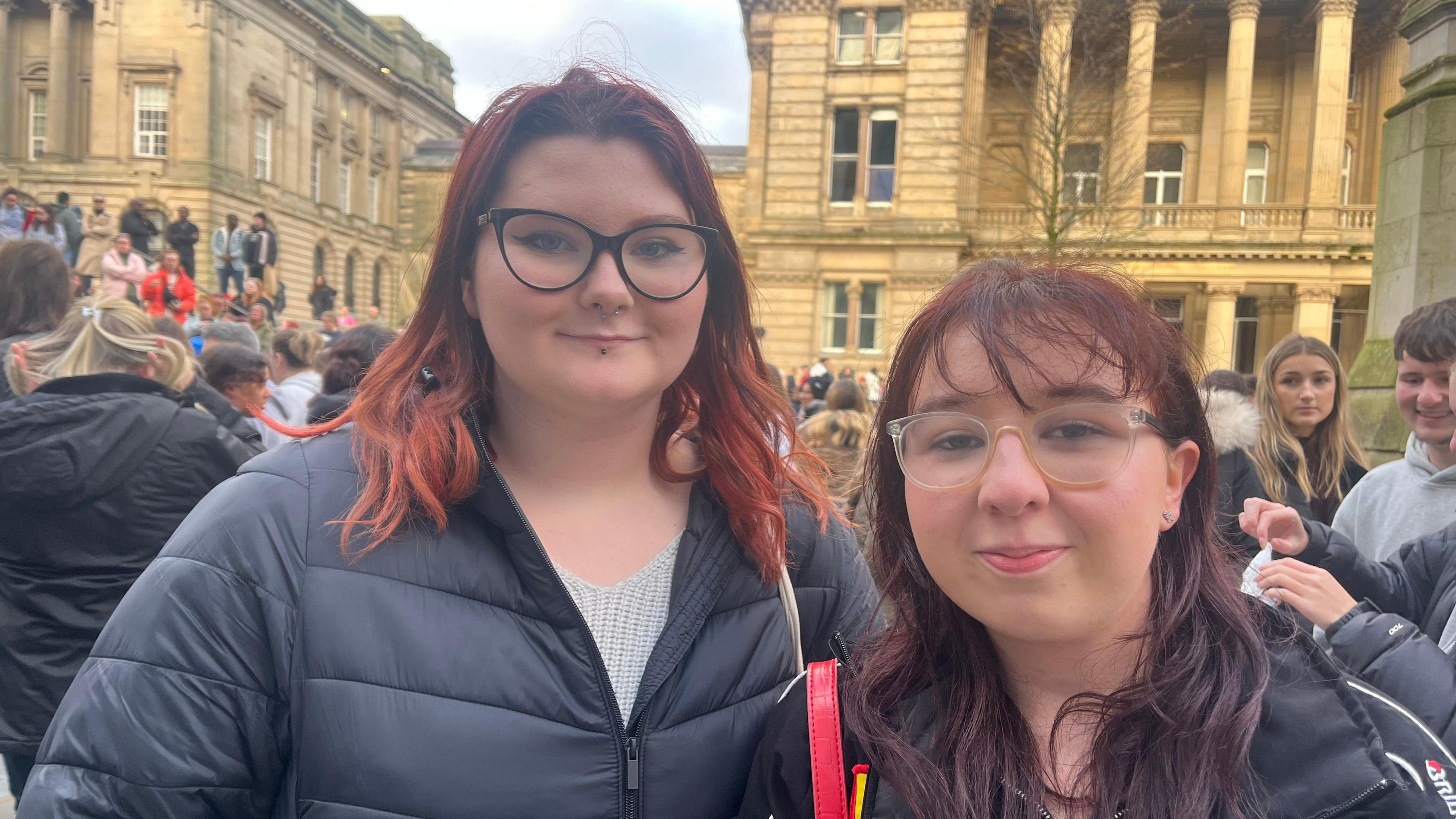 Two women with red hair stand together and smile at the camera. They are both wearing black coats, while the woman on the left has black glasses and the woman on the right has clear-coloured glasses. They stand in front of crowds of people gathered together, with buildings in the background.