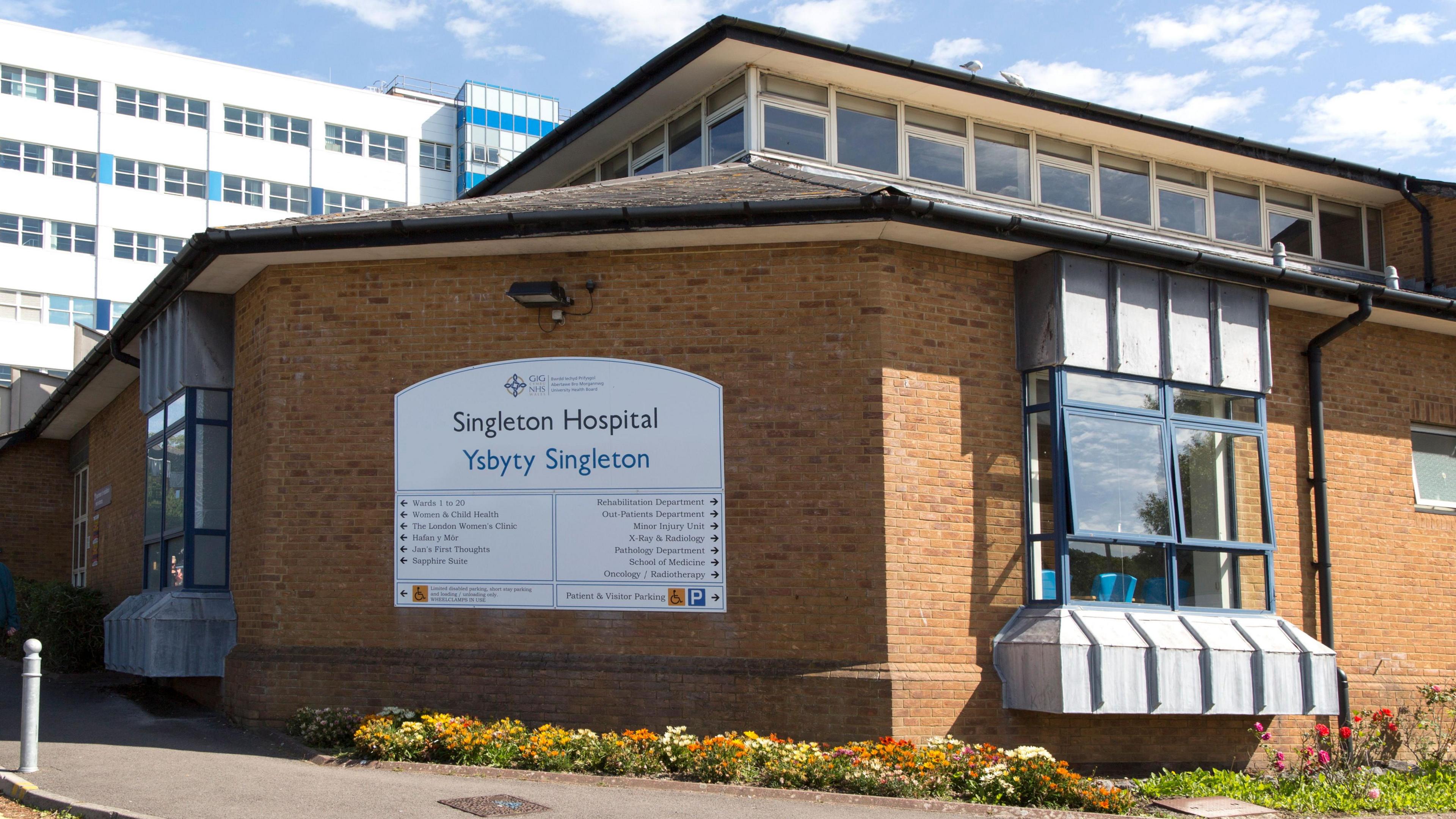A general view of Singleton Hospital. A single-storey brown brick building with a row of windows at the top has a large white sign on with the words 'Singleton Hospital'. Behind it is a large multi-storey white hospital building with rows of windows, with blue skies beyond.